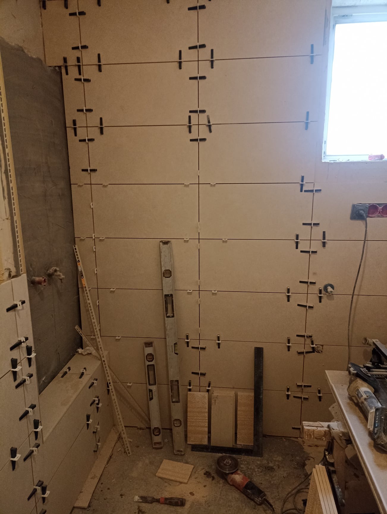 Ściana łazienki w trakcie układania płytek ceramicznych z użyciem poziomnic i klipsów dystansowych, widoczne narzędzia i materiały budowlane.