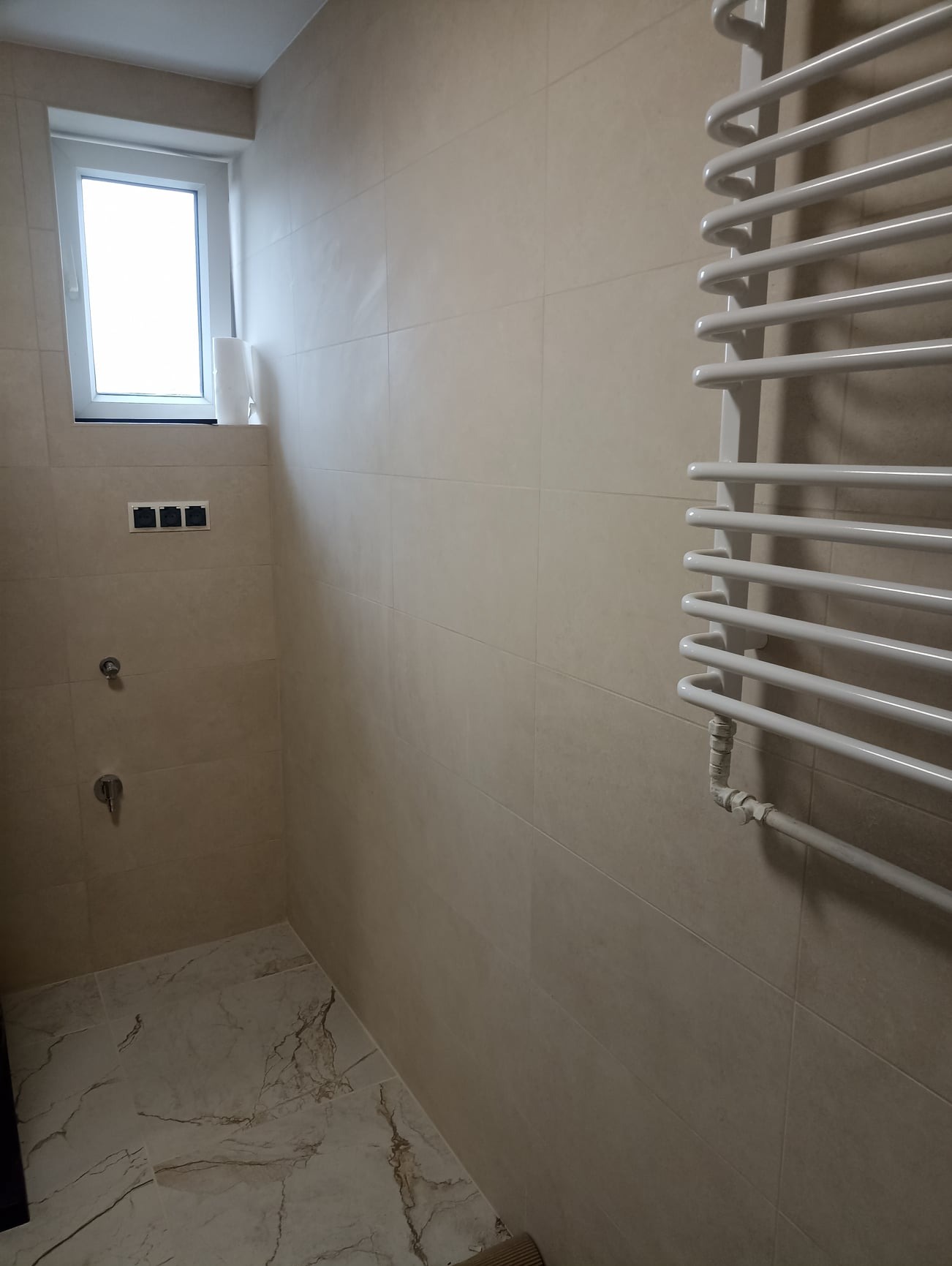 Wnętrze wykończonej łazienki z beżowymi płytkami, marmurową podłogą, białym grzejnikiem drabinkowym i oknem z widocznymi puszkami elektrycznymi.