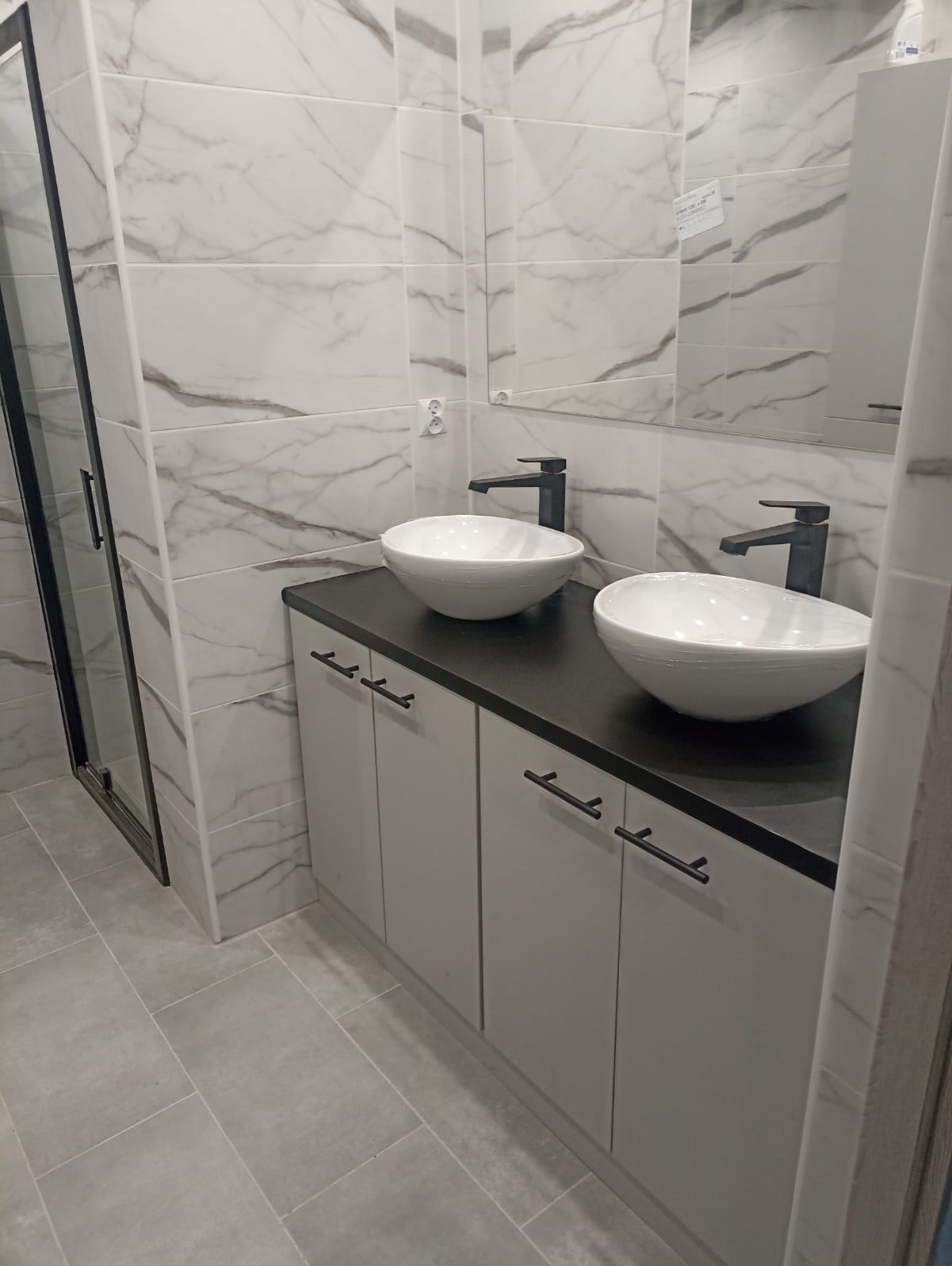 Nowoczesna łazienka z dwoma okrągłymi umywalkami nablatowymi, czarną armaturą i szarymi szafkami pod umywalką, wykończona płytkami imitującymi marmur.