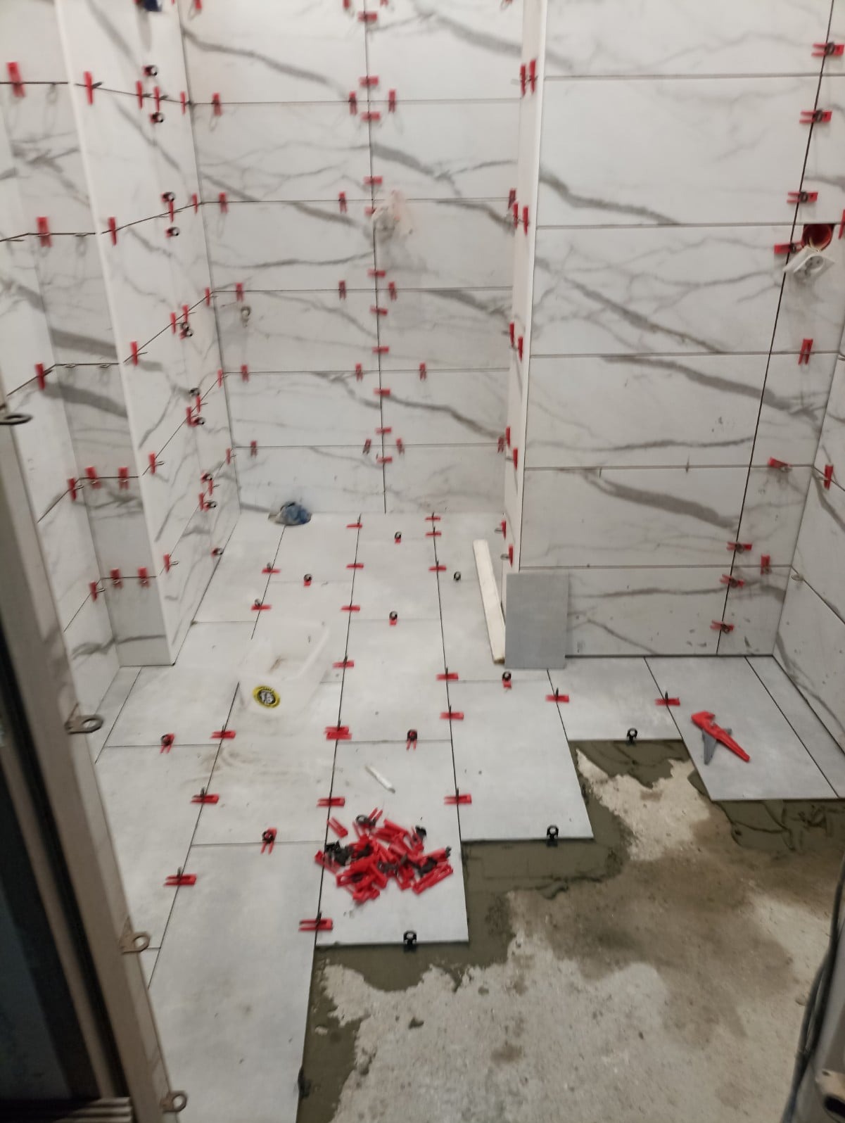 Wnętrze łazienki w trakcie układania płytek imitujących marmur, widoczne czerwone klipsy poziomujące, sznurki traserskie i częściowo ułożona podłoga.