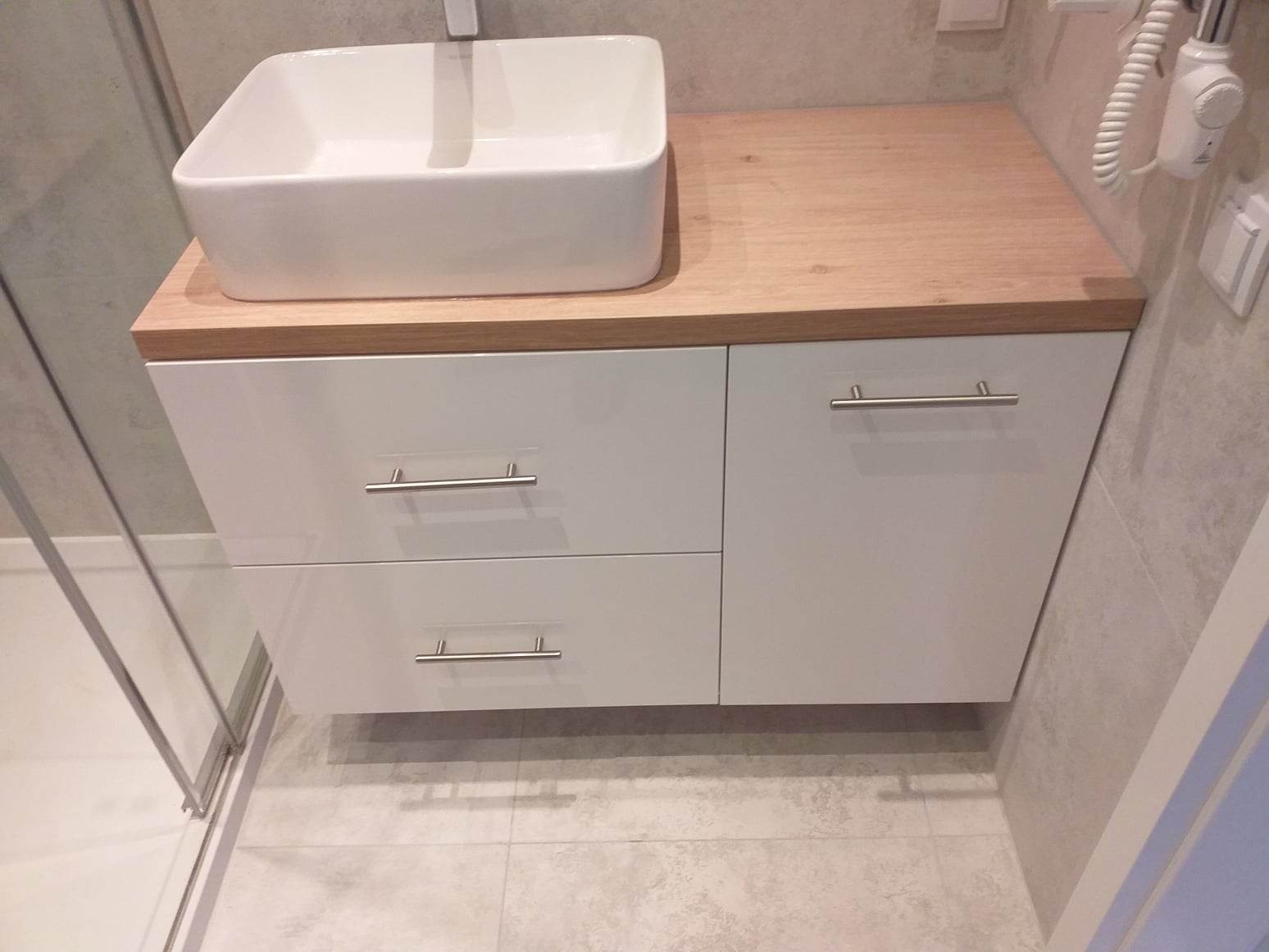 Nowoczesna biała szafka łazienkowa z umywalką nablatową i drewnianym blatem, minimalistyczne uchwyty, szare płytki podłogowe i fragment kabiny prysznicowej.