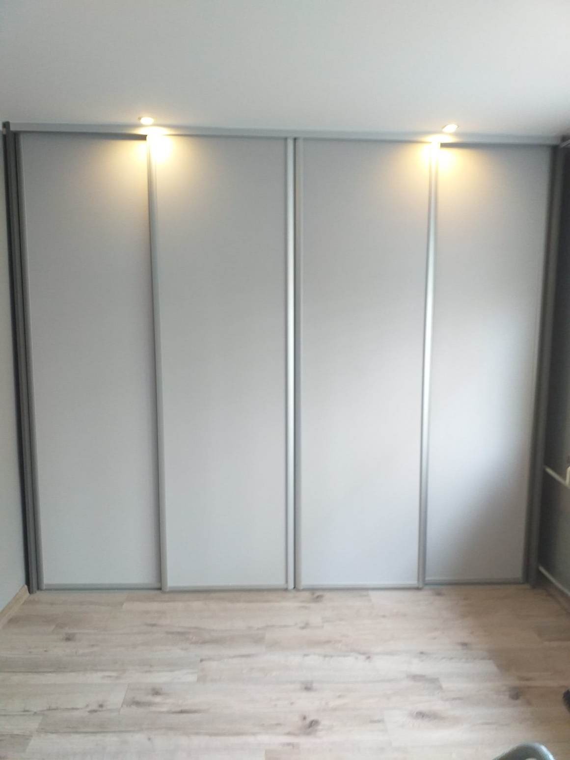 Szara szafa wnękowa z przesuwnymi drzwiami i oświetleniem punktowym w mieszkaniu w Krakowie, widok na podłogę z paneli imitujących drewno.