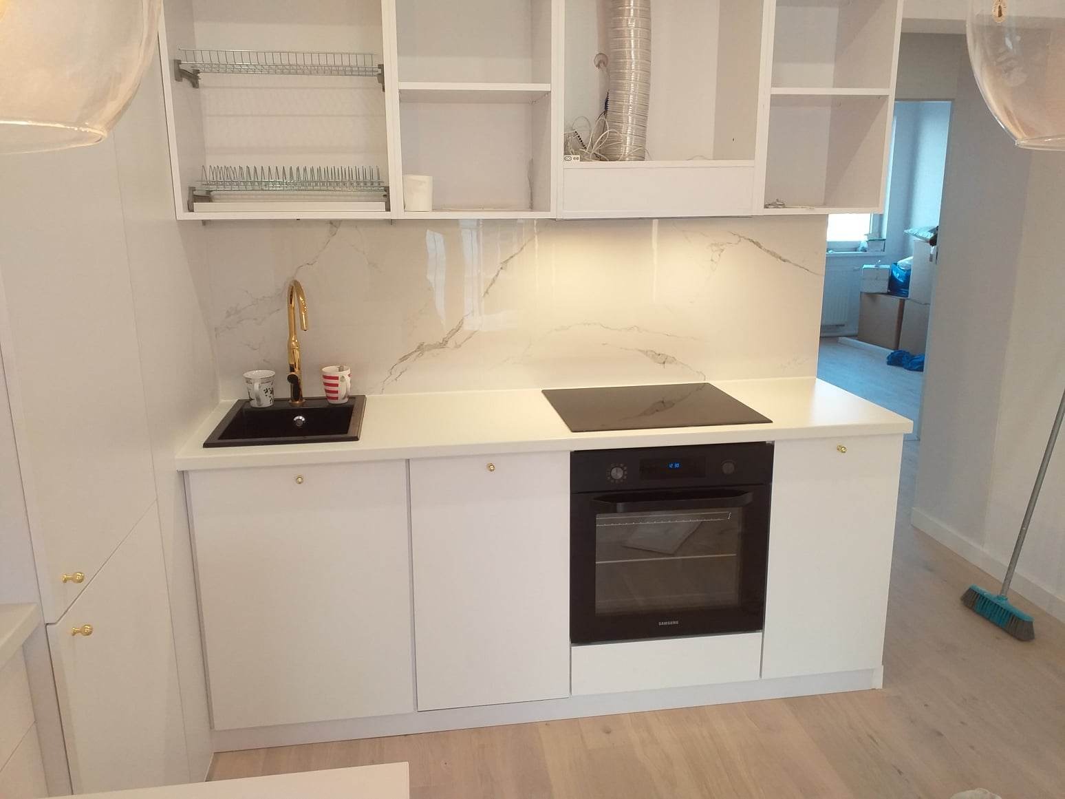 Nowoczesna kuchnia z białymi, lakierowanymi frontami, złotą baterią i marmurowym panelem ściennym, wyposażona w płytę indukcyjną i piekarnik Samsung.