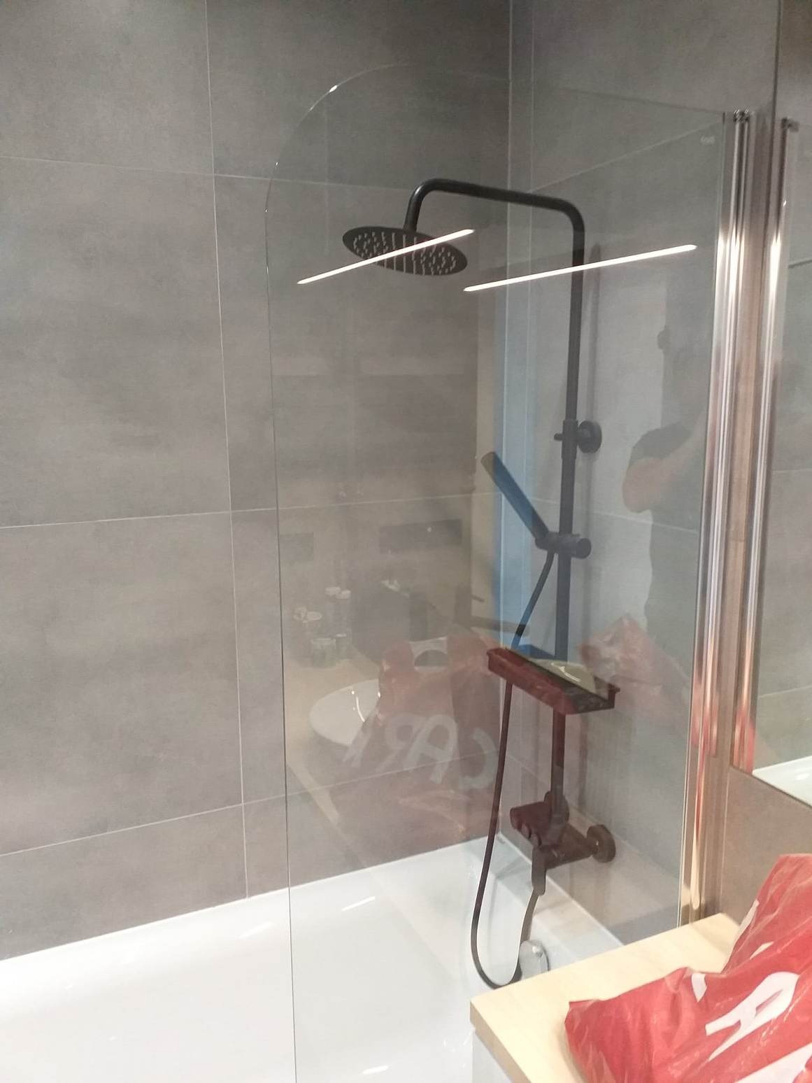 Nowoczesna kabina prysznicowa z czarną armaturą i szklaną ścianką, widoczne szare płytki na ścianie, biała wanna i element blatu z czerwonym workiem w rogu.