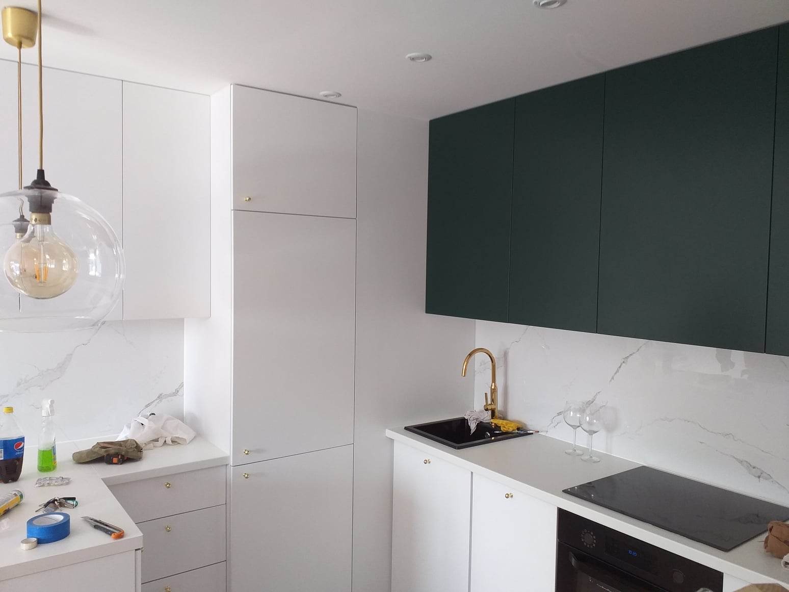 Nowoczesna kuchnia z białymi szafkami dolnymi i wysokimi, ciemnozielonymi szafkami górnymi, złotą armaturą, marmurowym backsplashiem i wiszącą lampą z dekoracyjną żarówką.
