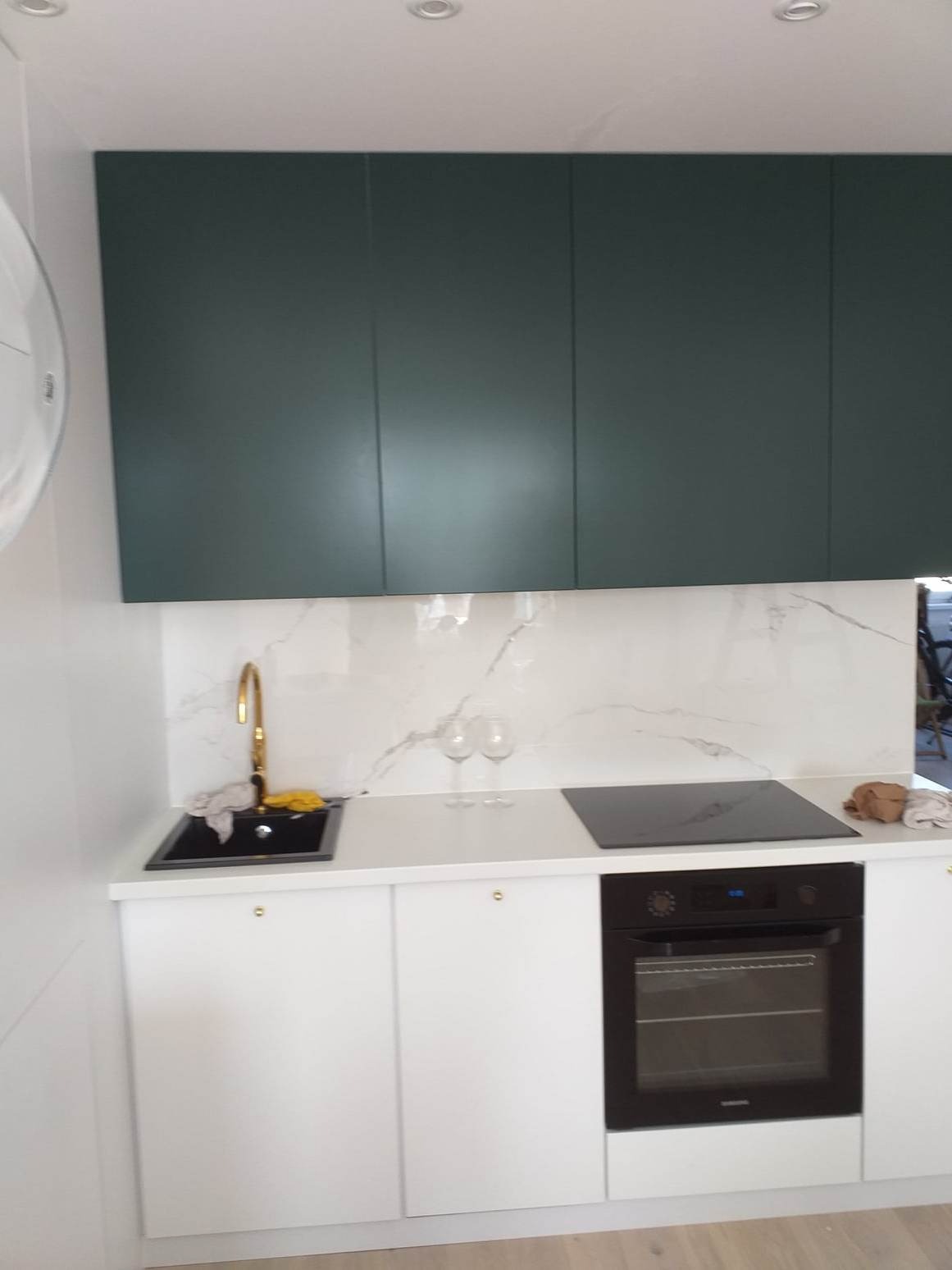 Nowoczesna kuchnia z ciemnozielonymi szafkami górnymi, białym blatem z marmurowym wzorem i złotą baterią, czarny zlew i płyta indukcyjna.