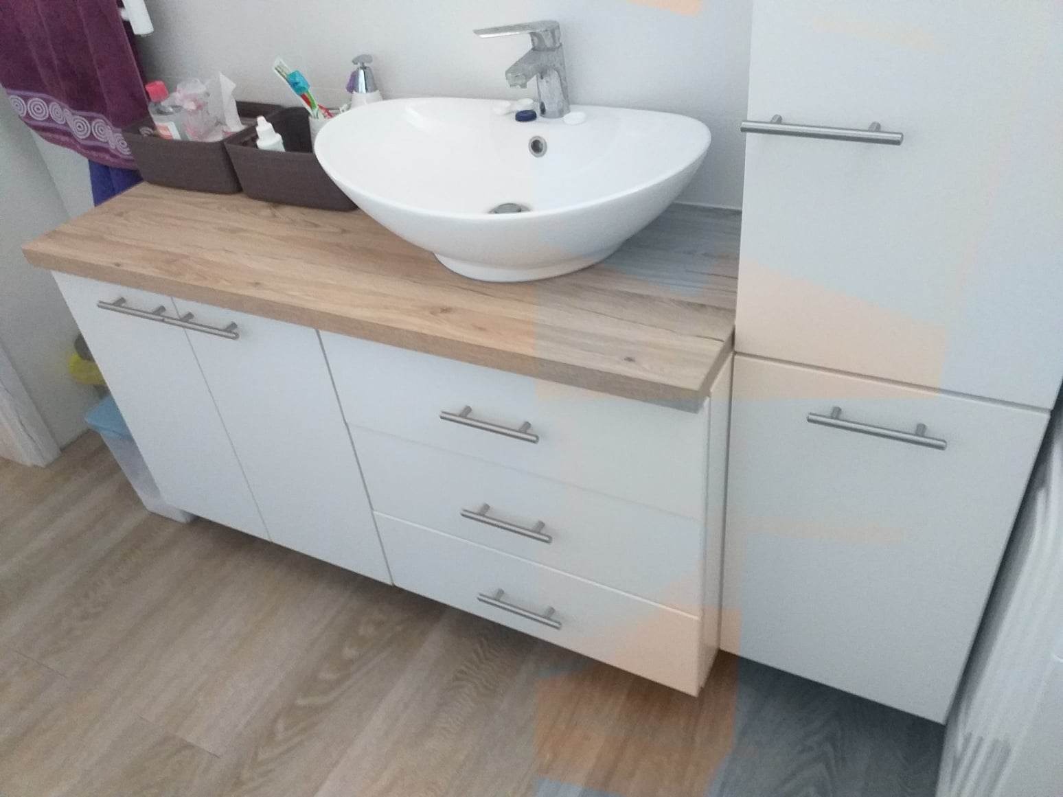 Nowoczesna umywalka nablatowa w kształcie misy, osadzona na blacie imitującym jasne drewno, pod nią białe szafki z trzema szufladami i drzwiczkami, całość w łazience wykończonej panelami...