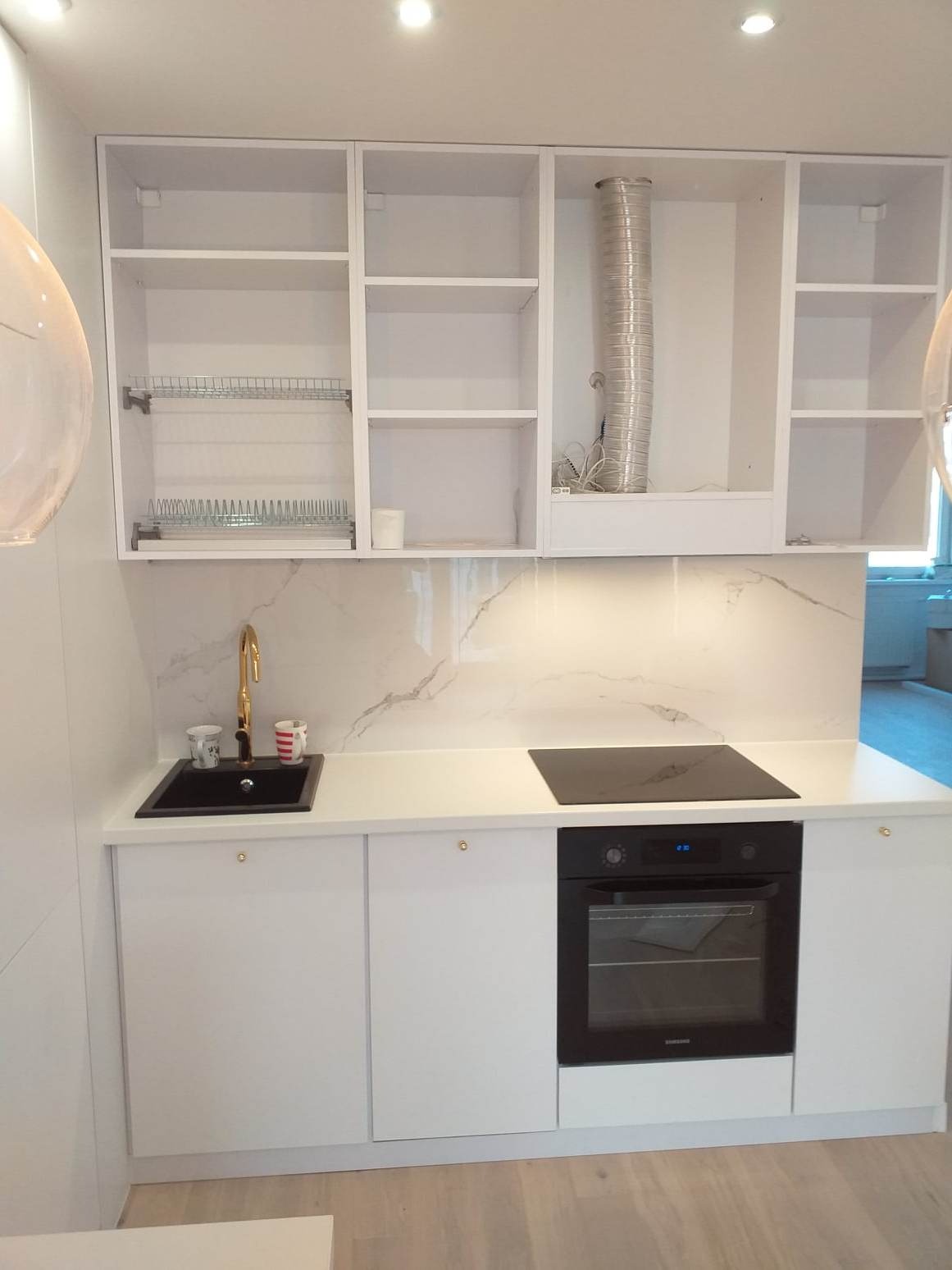 Jasna kuchnia z białymi szafkami, czarnym zlewem i złotą baterią, marmurowym backsplashiem i płytą indukcyjną, widoczne akcesoria kuchenne.