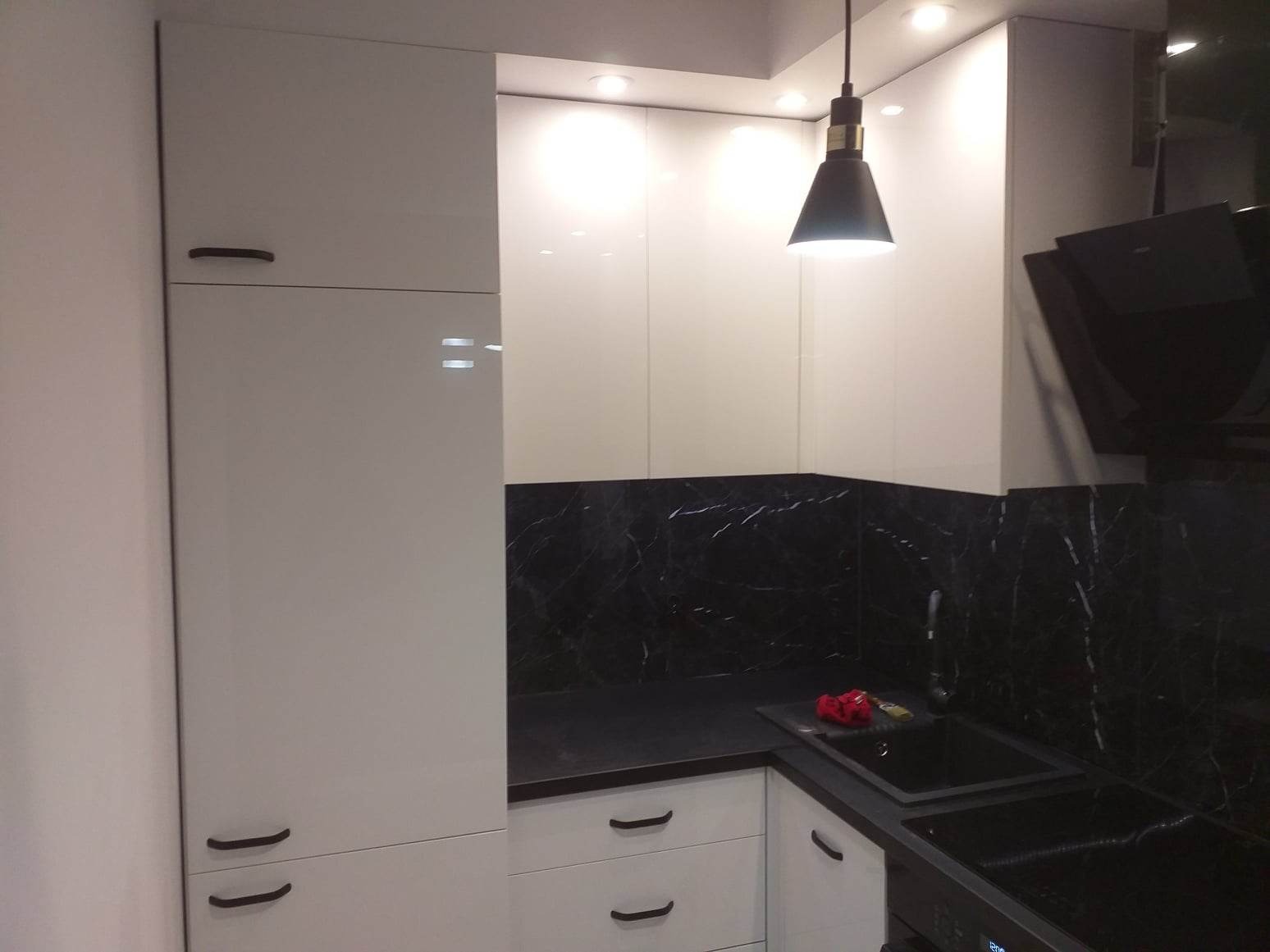 Nowoczesna kuchnia z białymi szafkami na wysoki połysk, czarnym blatem i marmurowym panelem ściennym, oświetlona wiszącą lampą z czarnym kloszem.