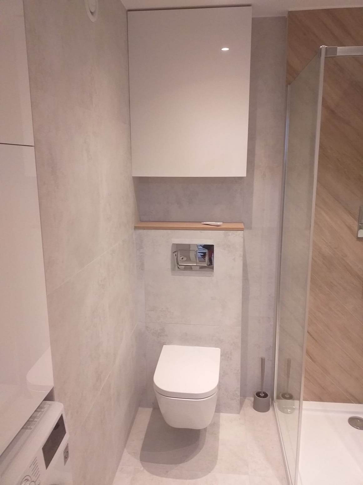 Nowoczesna łazienka z wiszącą toaletą, szarymi płytkami imitującymi beton, białymi szafkami i kabiną prysznicową z drewnopodobnymi panelami na ścianie, widoczna szczotka do WC.