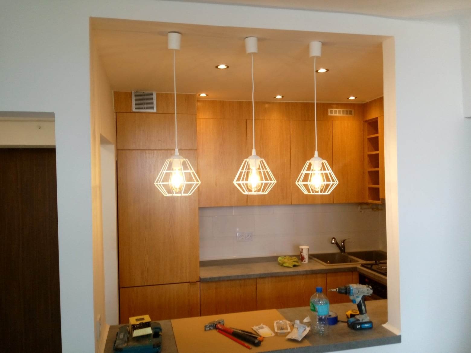 Nowoczesna kuchnia z drewnianymi szafkami i trzema identycznymi lampami wiszącymi o geometrycznym kształcie, na blacie widoczne narzędzia i materiały budowlane.