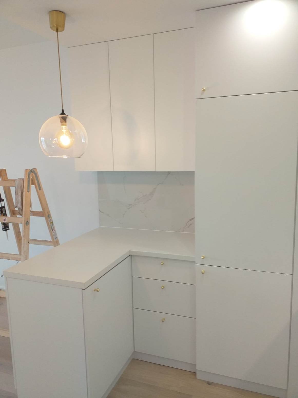 Biała zabudowa kuchenna z marmurowym backsplashiem i złotymi gałkami, widoczna drabina malarska sugeruje trwający remont.