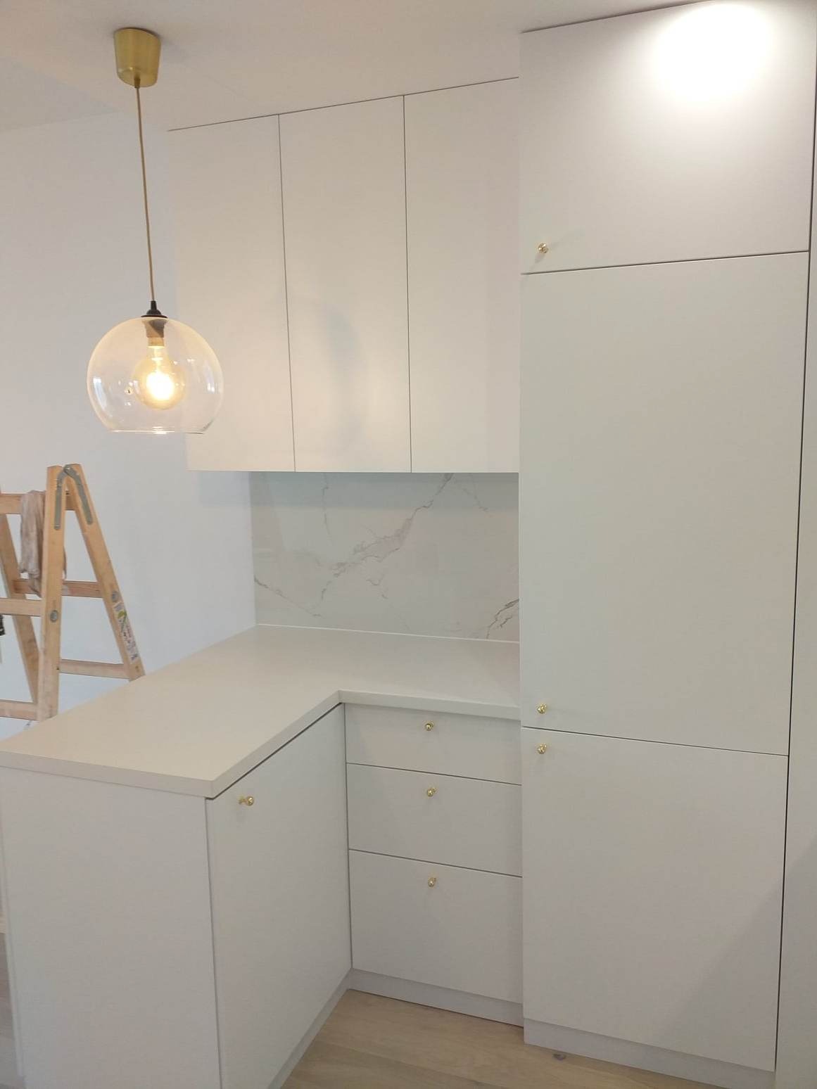 Nowoczesna kuchnia z białymi, matowymi frontami szafek, złotymi uchwytami i blatem z jasnego konglomeratu, oświetlona wiszącą lampą ze szklanym kloszem, z widoczną drabiną malarską w tle.