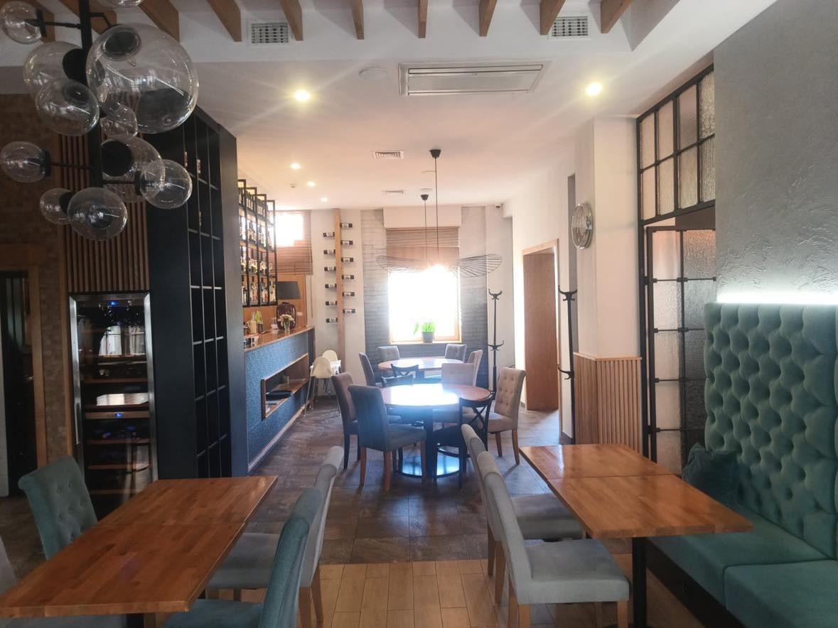 Wnętrze restauracji z drewnianymi stołami, krzesłami tapicerowanymi w odcieniach szarości i błękitu oraz dekoracyjnymi lampami wiszącymi z przezroczystego szkła. W tle bar z ekspozycją butelek.