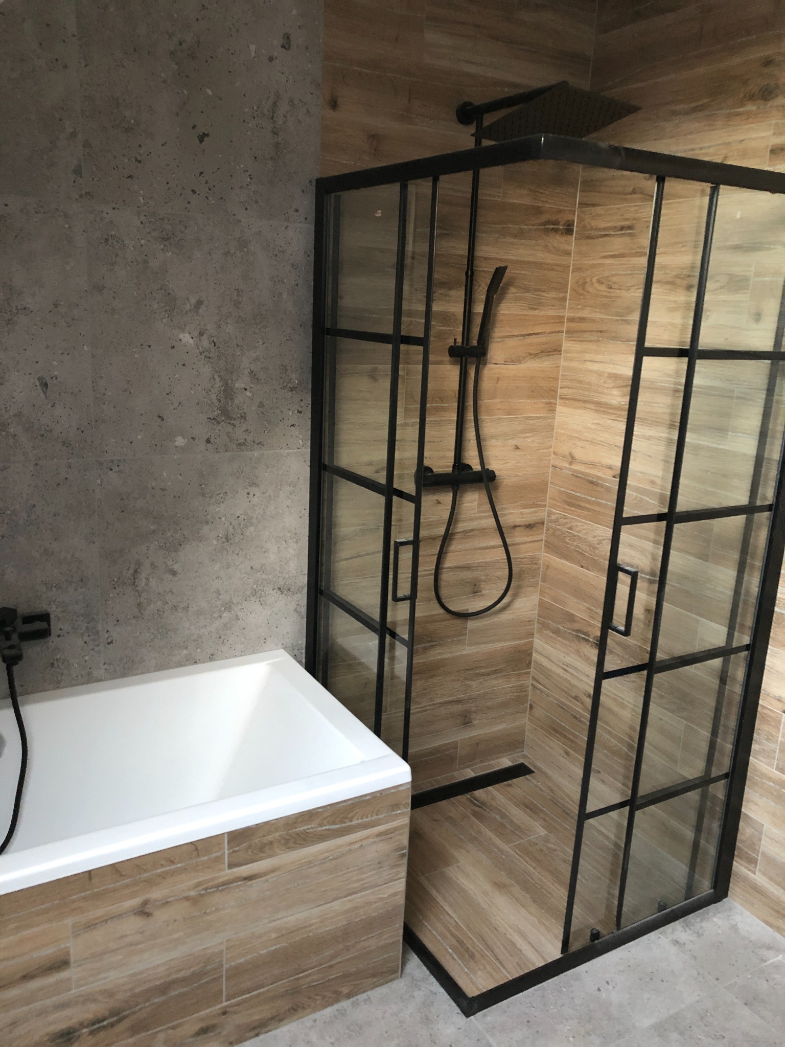 Nowoczesna łazienka z kabiną prysznicową typu walk-in z czarnymi profilami i deszczownicą, obok wanna obudowana płytkami imitującymi drewno, ściana wykończona szarym betonem architektonicznym.