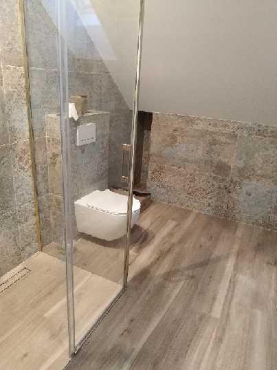 Nowoczesna łazienka w odcieniach beżu i szarości z kabiną prysznicową typu walk-in, wiszącą toaletą i płytkami imitującymi drewno na podłodze i ścianach.