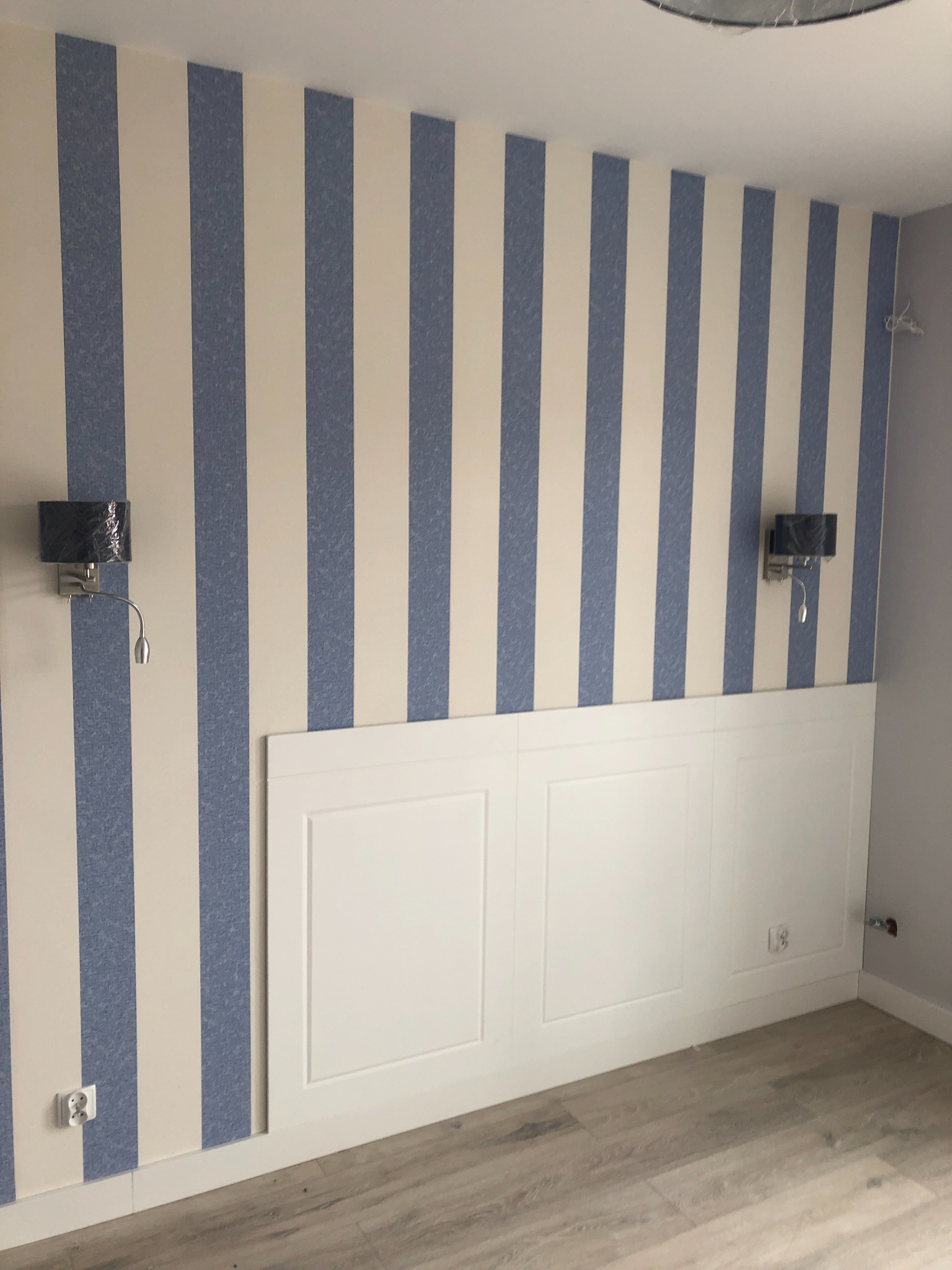 Pokój po odświeżeniu: ściany w biało-niebieskie pasy, biała boazeria panelowa do połowy wysokości, dwie czarne lampki nocne z regulowanymi ramionami, panele podłogowe w odcieniach szarości.