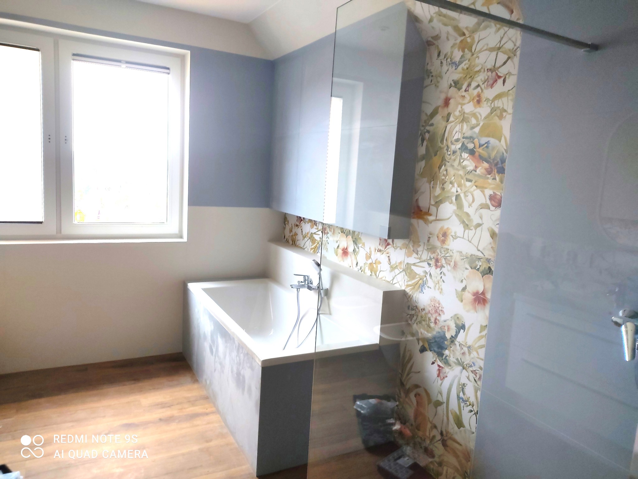 Nowoczesna łazienka z wanną obudowaną szarym betonem architektonicznym, szafkami w kolorze gołębim i tapetą w kwiaty i ptaki, widok na kabinę prysznicową ze szklanymi drzwiami.