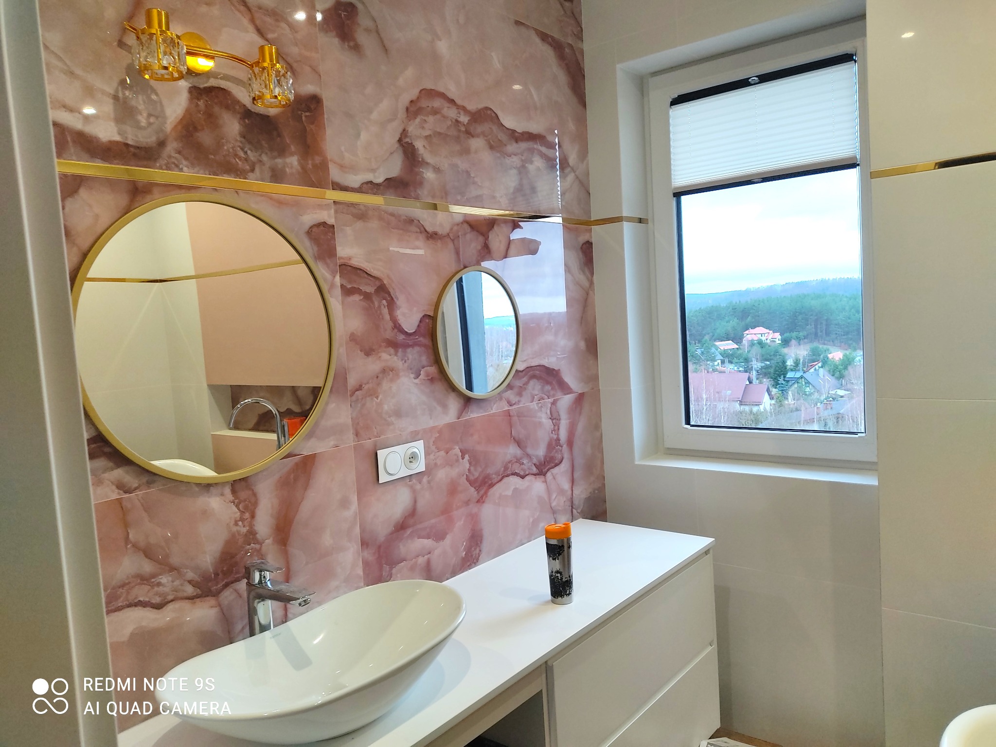 Wykończona łazienka z marmurowymi płytkami w odcieniach różu i złota, owalną umywalką nablatową, dwoma okrągłymi lustrami w złotych ramach i widokiem na las za oknem.