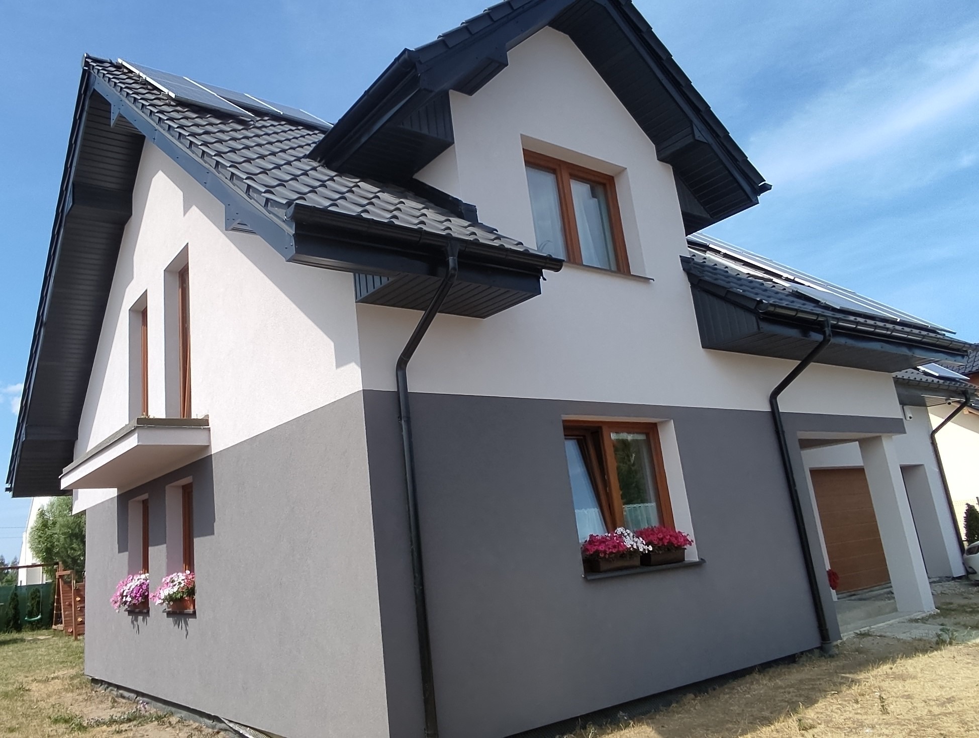Dwukolorowa elewacja domu jednorodzinnego z ciemną dachówką i panelami słonecznymi, okna z kwiatami w skrzynkach.