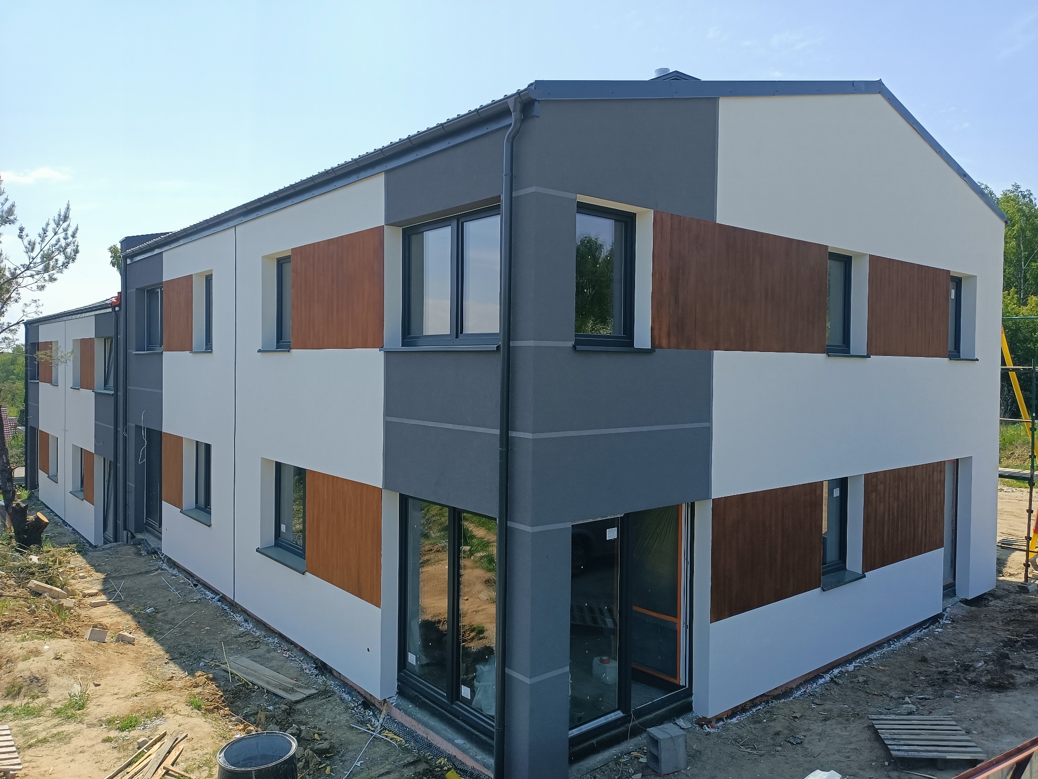 Nowoczesny, dwukondygnacyjny dom z elewacją w odcieniach szarości, bieli i brązu, widoczny z perspektywy lotu ptaka, z odsłoniętym gruntem wokół fundamentów i rusztowaniem w tle.