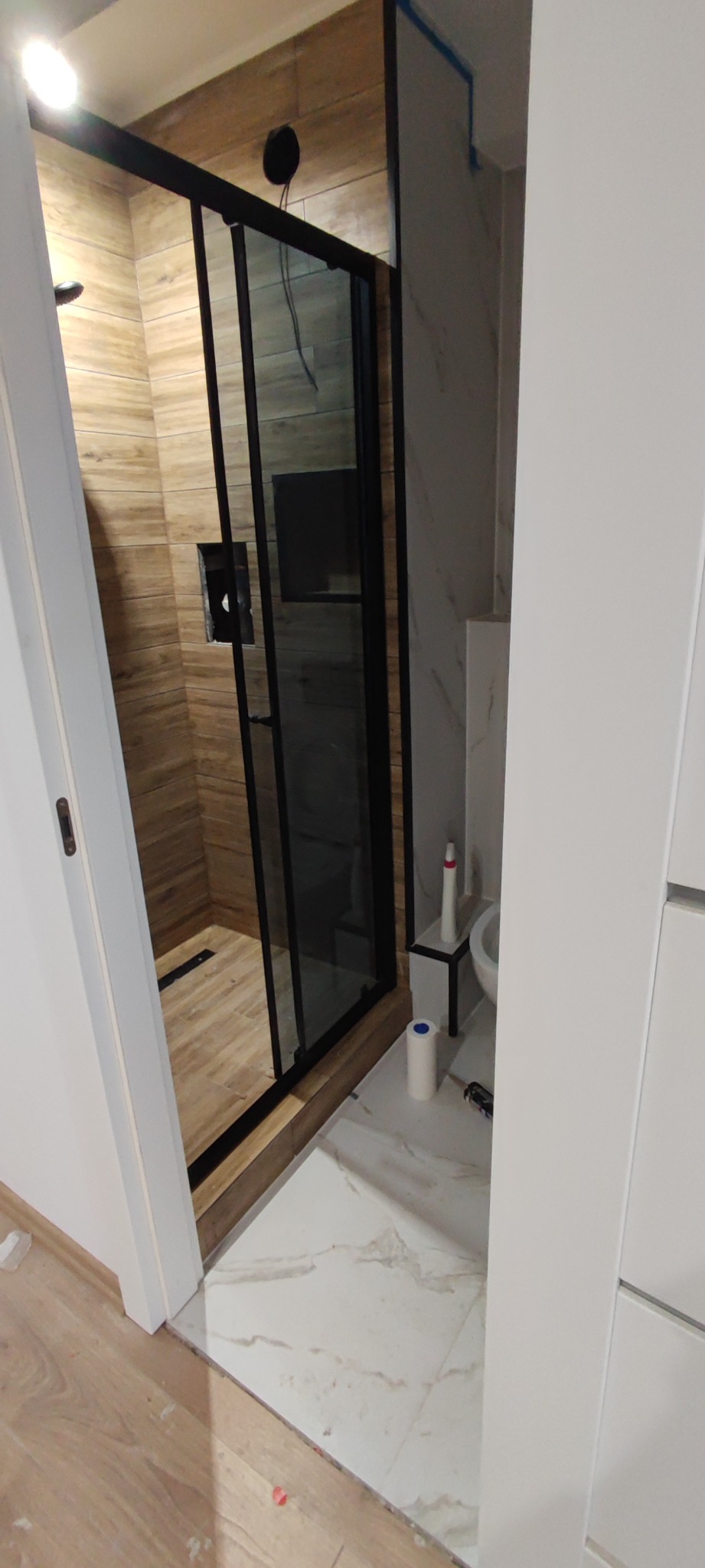 Nowoczesna kabina prysznicowa z czarnymi profilami i drewnopodobnymi płytkami wewnątrz, widok z zewnątrz na marmurowe płytki podłogowe i częściowo widoczną umywalkę z kosmetykami.