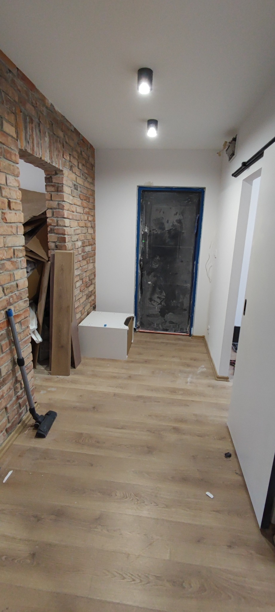 Korytarz w trakcie prac wykończeniowych: widoczna ceglana ściana z przejściem, surowa, zabezpieczona folią czarna ościeżnica drzwi, świeżo położona podłoga laminowana, biała szafka i odkurzacz przy...