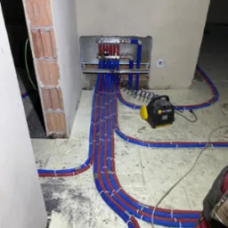 Rozdzielacz ogrzewania podłogowego z podłączonymi rurami w kolorze niebieskim i czerwonym, ułożonymi na podłodze w trakcie instalacji, widoczny agregat prądotwórczy i skrzynki narzędziowe.