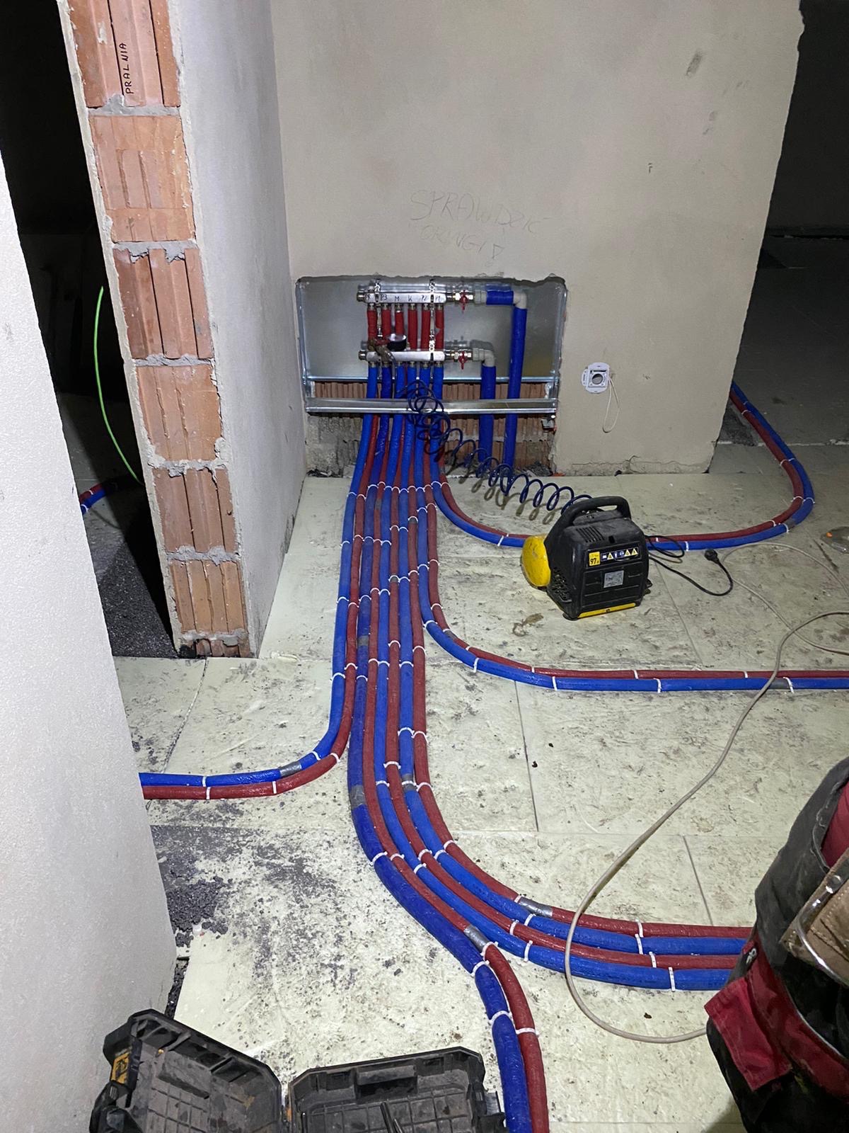 Rozdzielacz ogrzewania podłogowego z podłączonymi rurami w kolorze niebieskim i czerwonym, ułożonymi na podłodze w trakcie instalacji, widoczny agregat prądotwórczy i skrzynki narzędziowe.