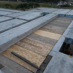 Technik - Montaż stropu Teriva z płyt OSB na budowie domu jednorodzinnego. Widoczne betonowe belki stropowe z okrągłymi otworami i drewniane płyty. Tło: zabudowa wiejska.