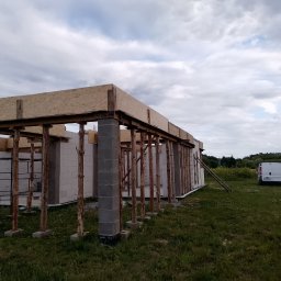 Technik - Wznoszenie ścian parterowego domu z betonu komórkowego. Widoczne podpory drewniane i szalunek stropu. Na zewnątrz trawa i zaparkowany biały bus.