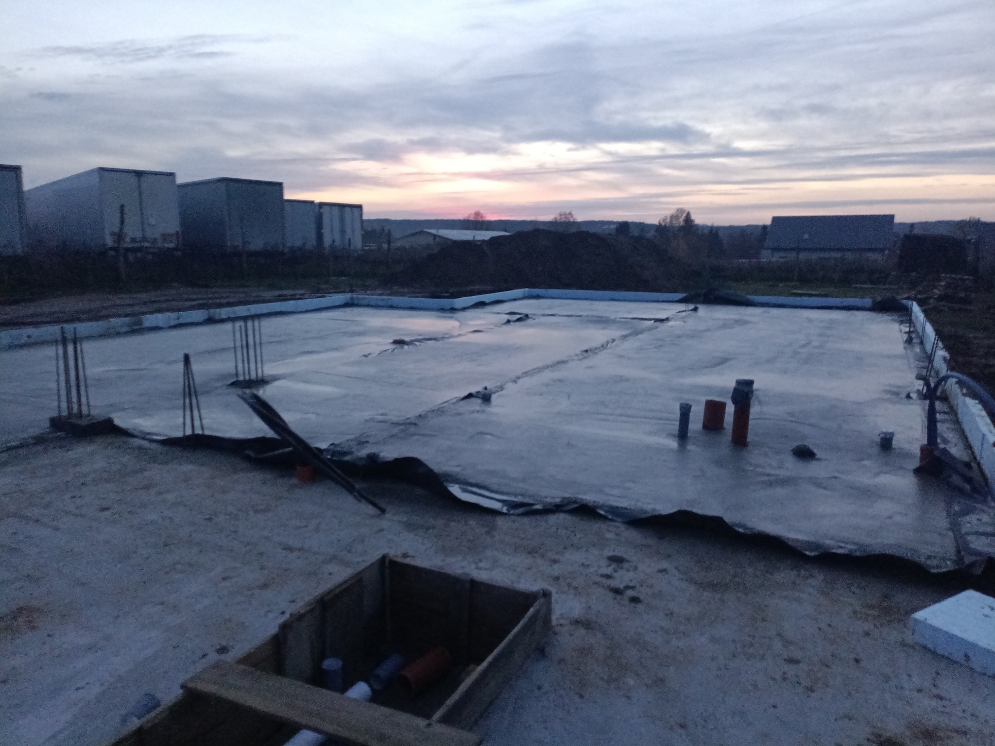 Zalana płyta fundamentowa z wystającymi prętami zbrojeniowymi i instalacją hydrauliczną na tle zachodzącego słońca i zabudowań gospodarczych.
