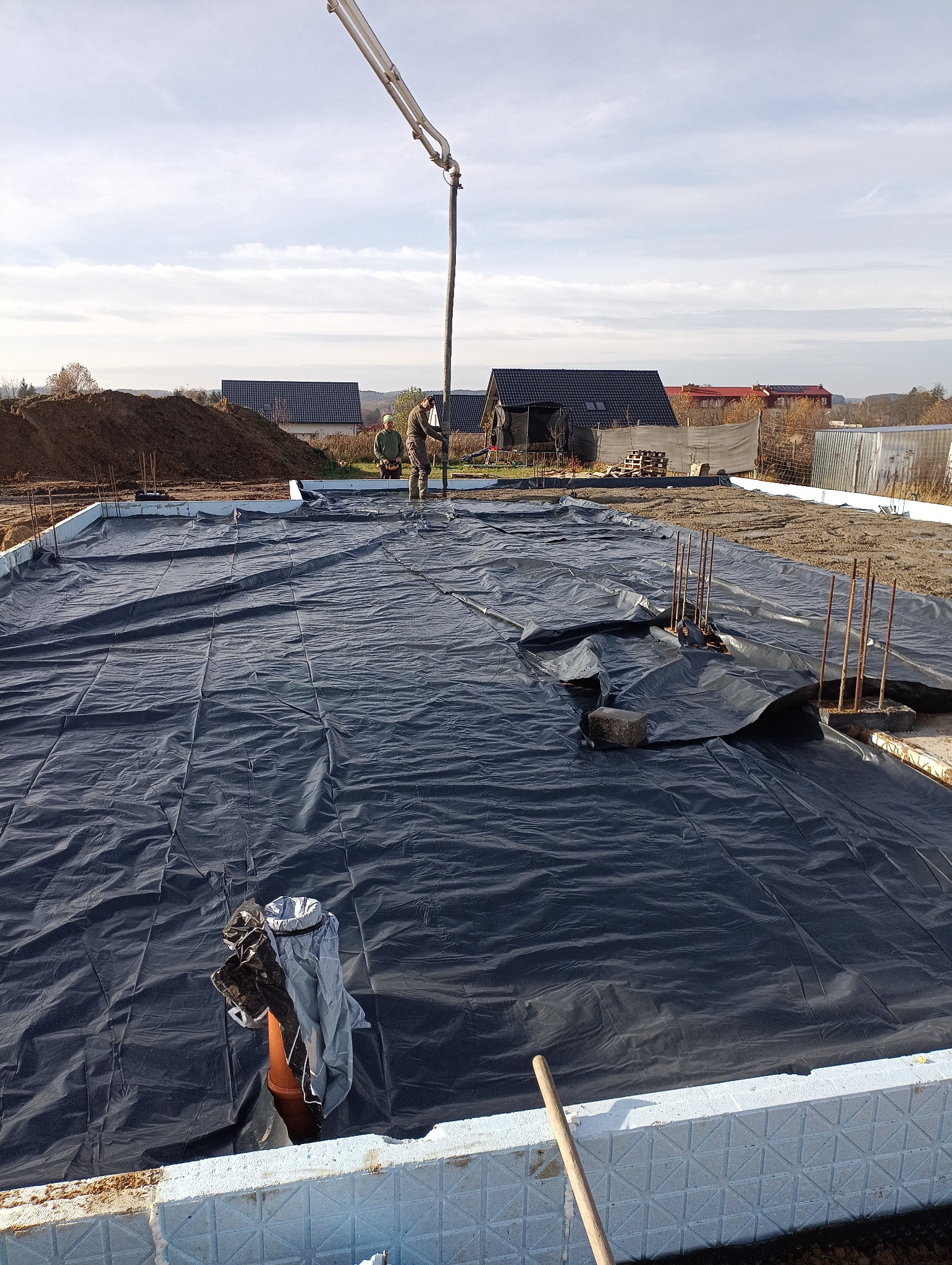 Wylewka fundamentów pod dom jednorodzinny. Widoczna izolacja termiczna, folia hydroizolacyjna i zbrojenie. W tle pracownicy obsługujący pompę do betonu.