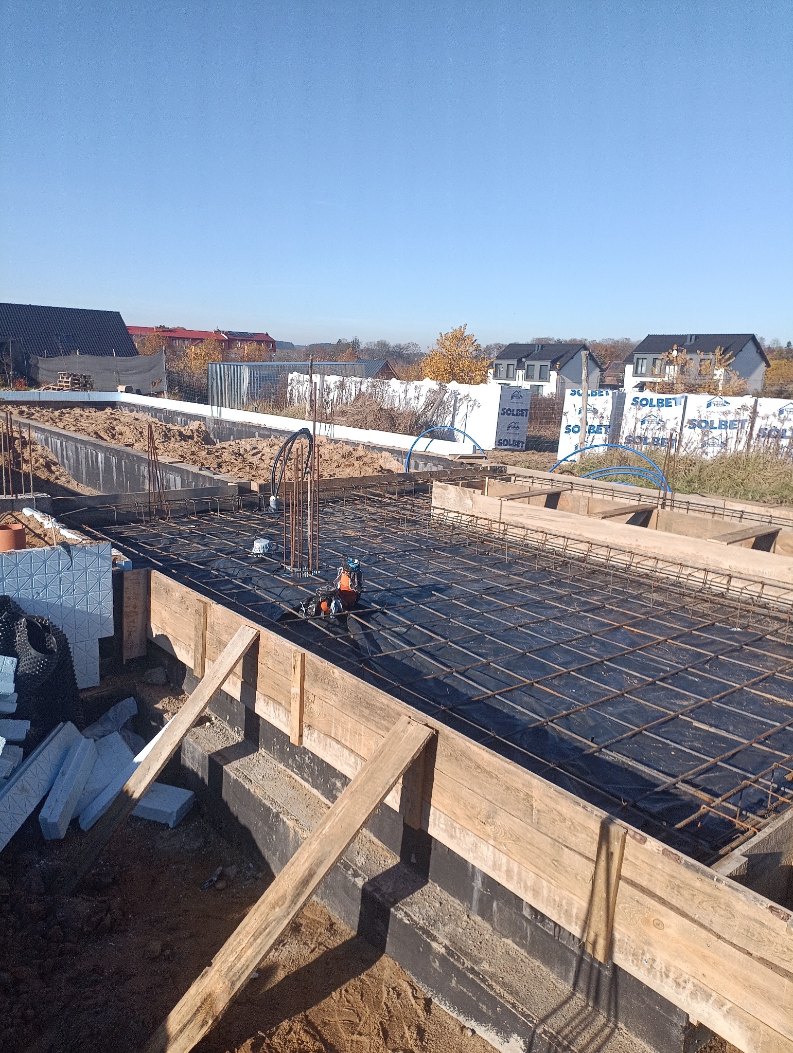 Zbrojenie fundamentów pod budowę domu jednorodzinnego. Widoczne szalunki, stalowe pręty zbrojeniowe i izolacja termiczna. W tle zabudowa jednorodzinna i błękitne niebo.