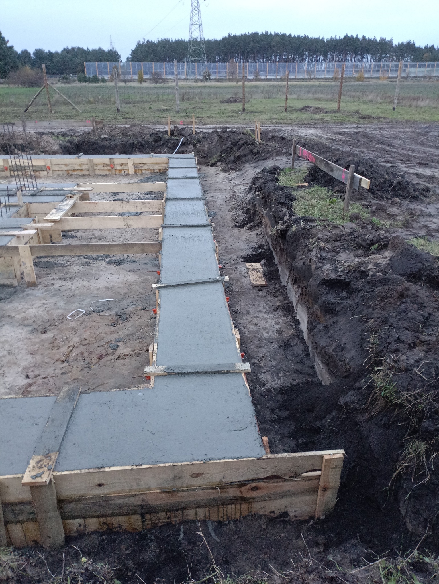 Zalewanie betonem szalunku fundamentów na budowie. Widoczne drewniane deski szalunkowe i świeży beton. W tle ziemia, trawa i słupy wysokiego napięcia.