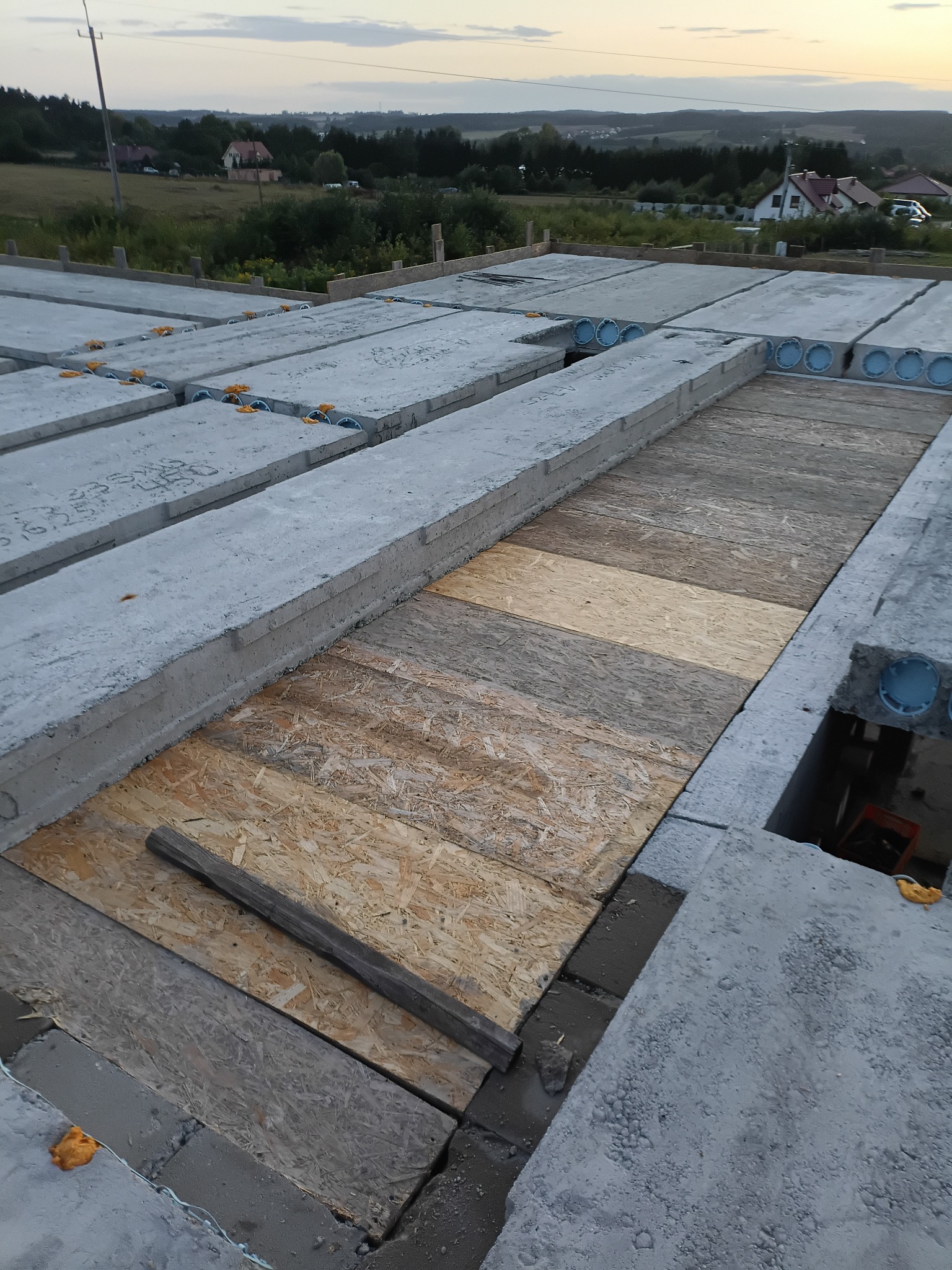 Montaż stropu Teriva z płyt OSB na budowie domu jednorodzinnego. Widoczne betonowe belki stropowe z okrągłymi otworami i drewniane płyty. Tło: zabudowa wiejska.