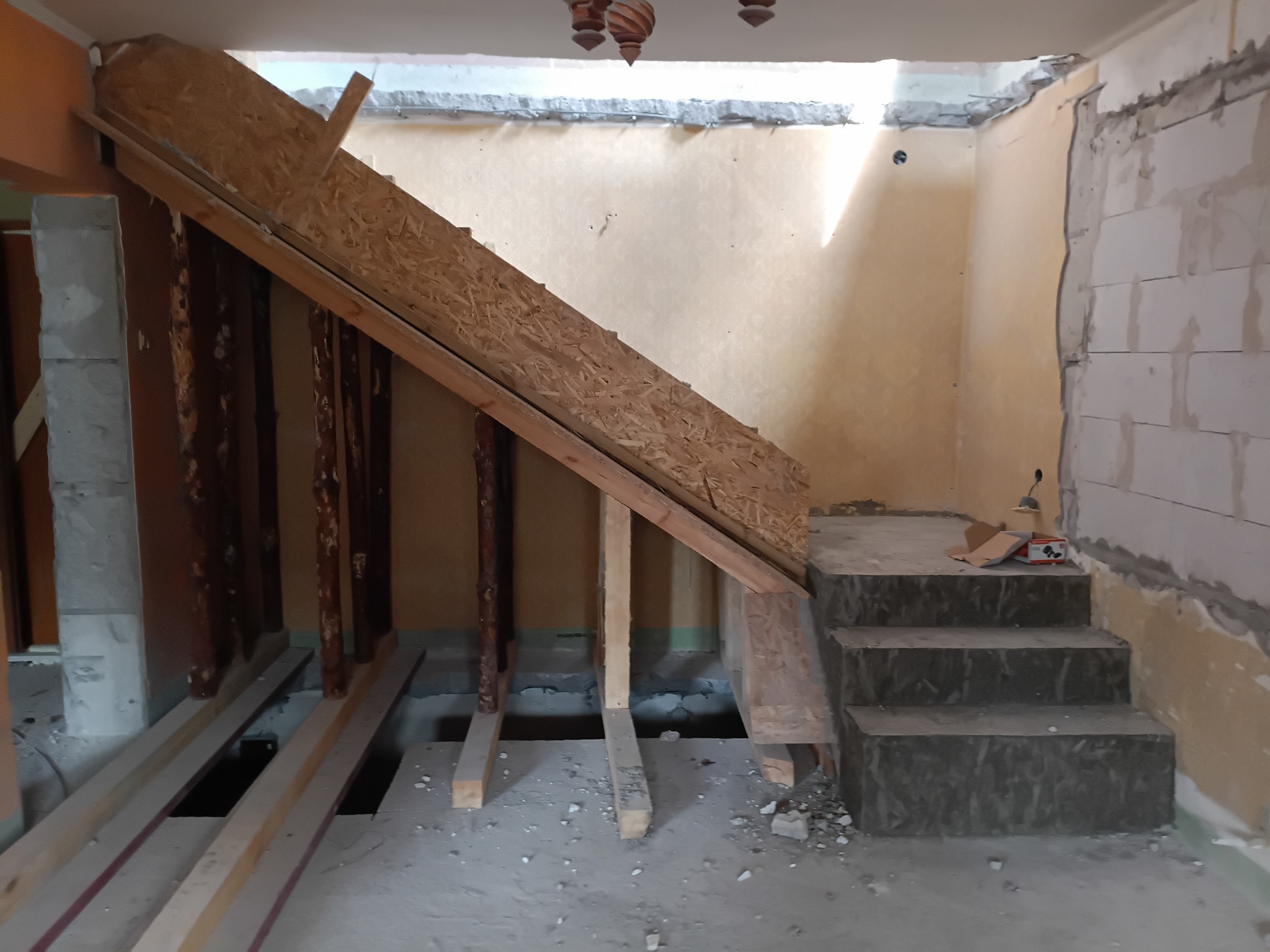 Wnętrze w trakcie remontu: drewniana konstrukcja schodów z płyt OSB, odsłonięte ściany z pustaków, przygotowane legary pod podłogę. Widoczne ślady prac budowlanych.