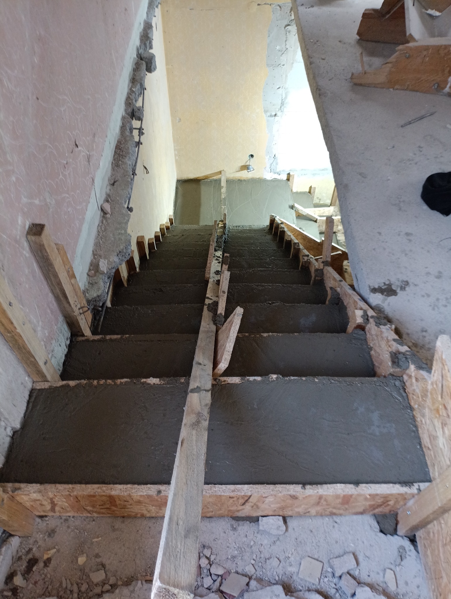 Wypełnione betonem schody w trakcie budowy z drewnianym szalunkiem, widok z góry. Surowe ściany w tle. Etap prac wykończeniowych.