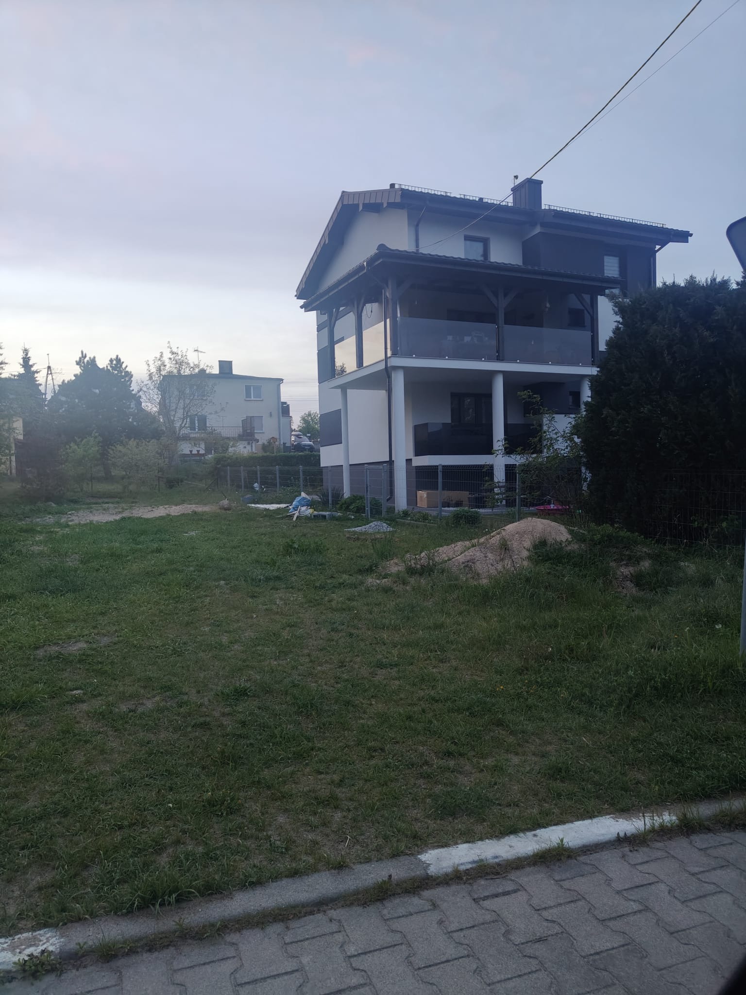 Dwupiętrowy dom z balkonami i szklanymi balustradami, widoczny z perspektywy ulicy, z niezagospodarowanym terenem przed budynkiem. Widoczne elementy wykończeniowe.