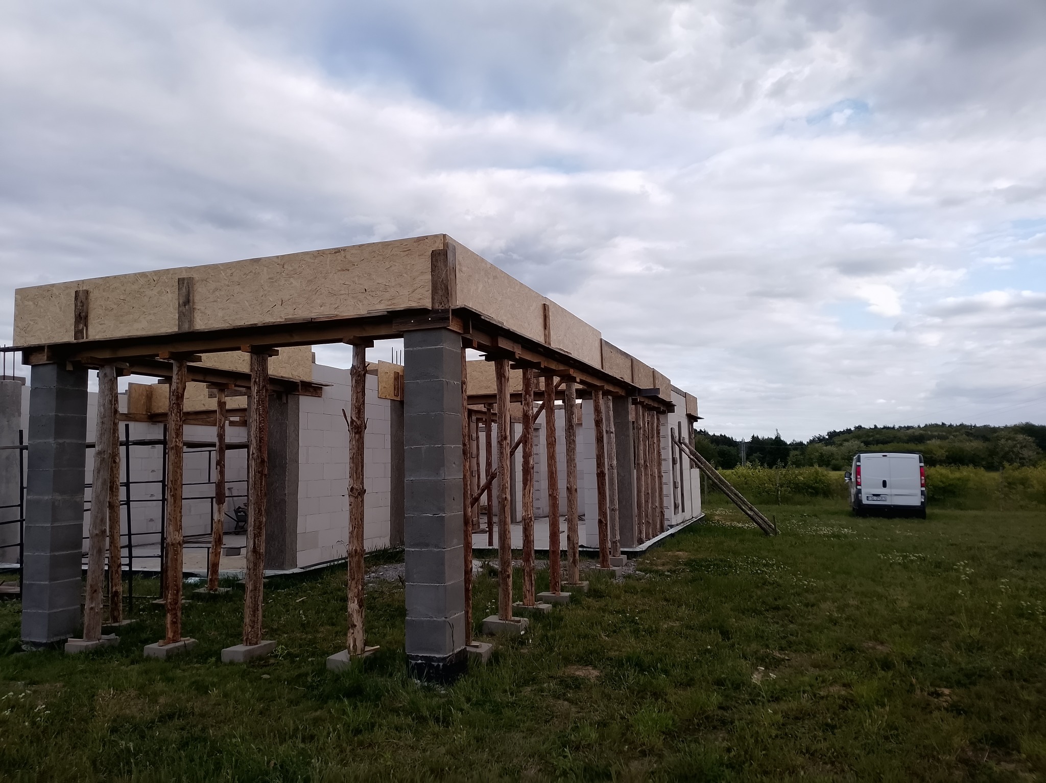 Wznoszenie ścian parterowego domu z betonu komórkowego. Widoczne podpory drewniane i szalunek stropu. Na zewnątrz trawa i zaparkowany biały bus.