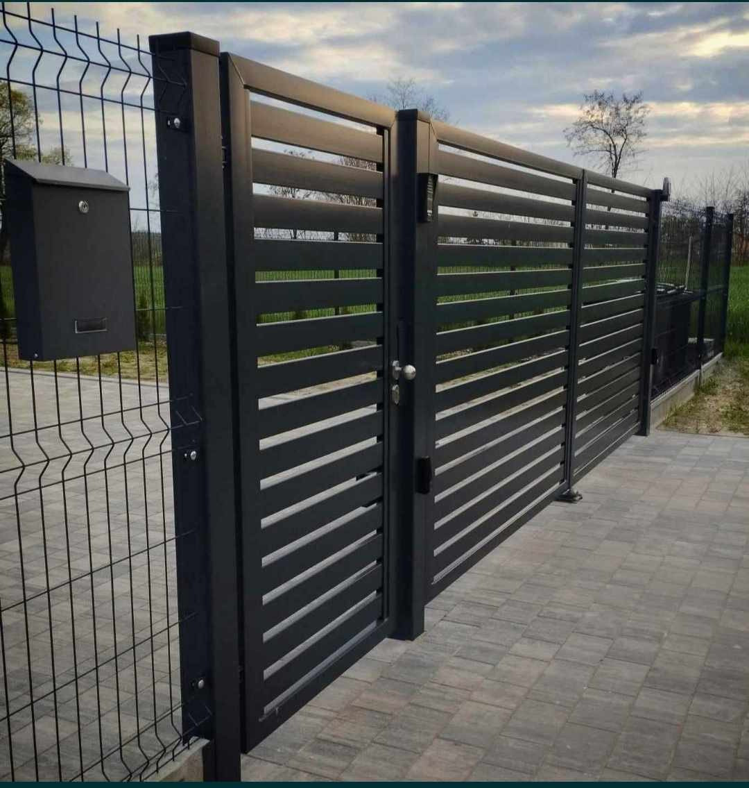 Nowoczesna, grafitowa brama wjazdowa z poziomych profili, przylegająca do ogrodzenia z siatki plecionej i skrzynki pocztowej, na tle zieleni.