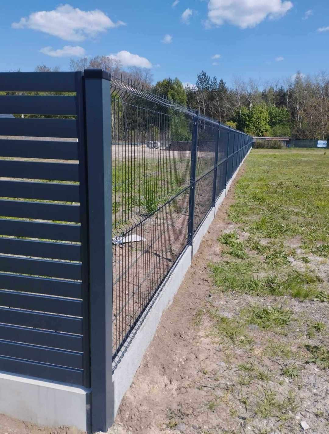 Długie, proste ogrodzenie z paneli i siatki na betonowej podmurówce, oddzielające trawnik od zalesionego terenu w tle.