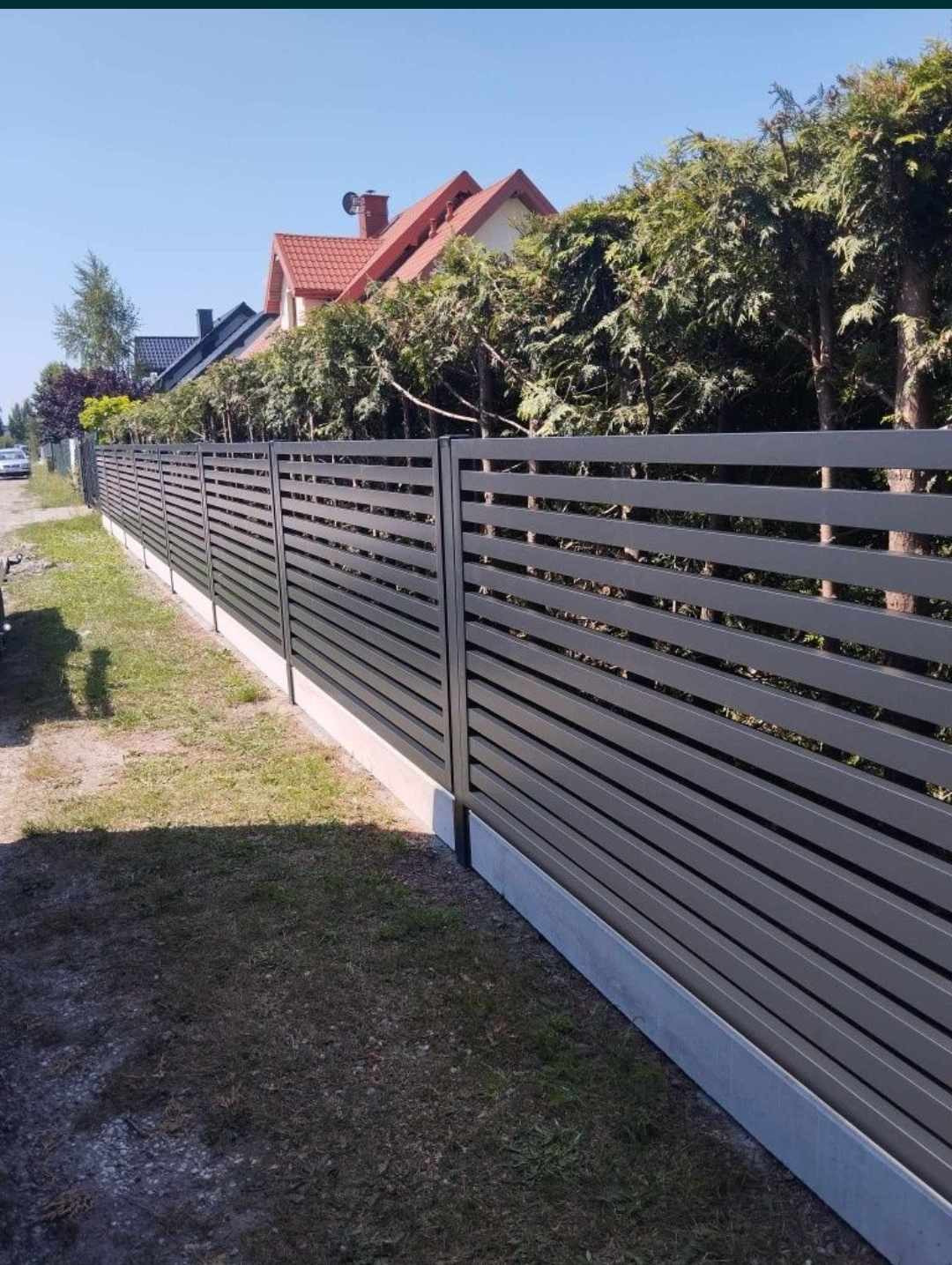 Długie, poziome, szare panele ogrodzeniowe z betnową podmurówką, oddzielające posesję od ulicy, z widocznymi domami w tle i zielonymi tujami wzdłuż ogrodzenia.
