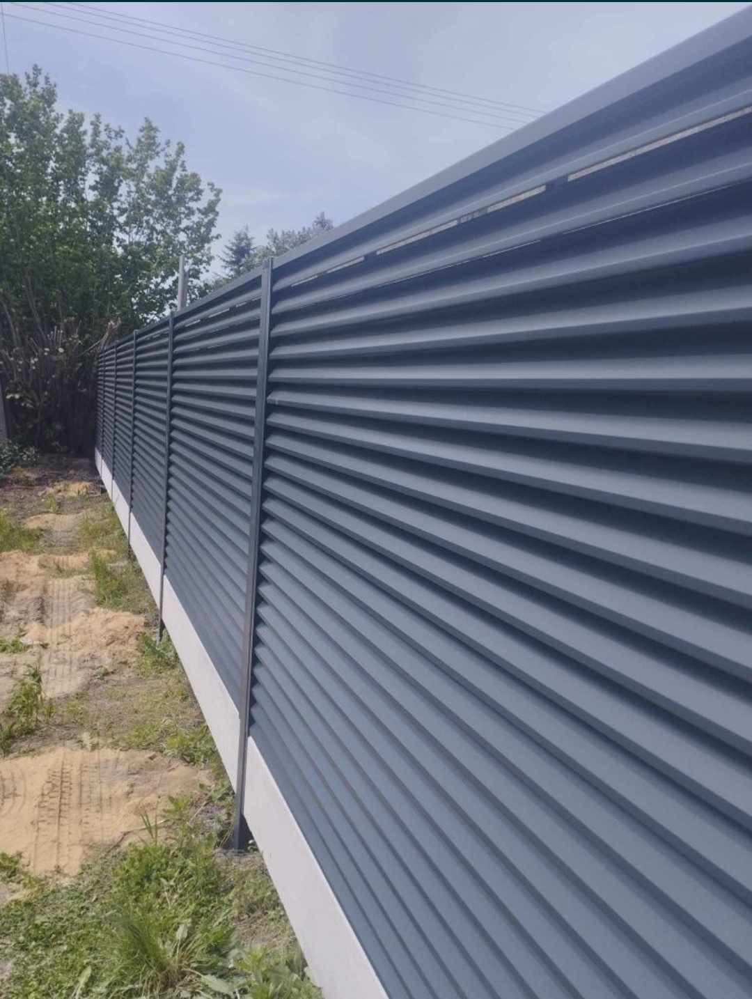 Nowoczesne ogrodzenie panelowe z poziomych, szarych profili, zamontowane na białej podmurówce, widoczne z boku w perspektywie.