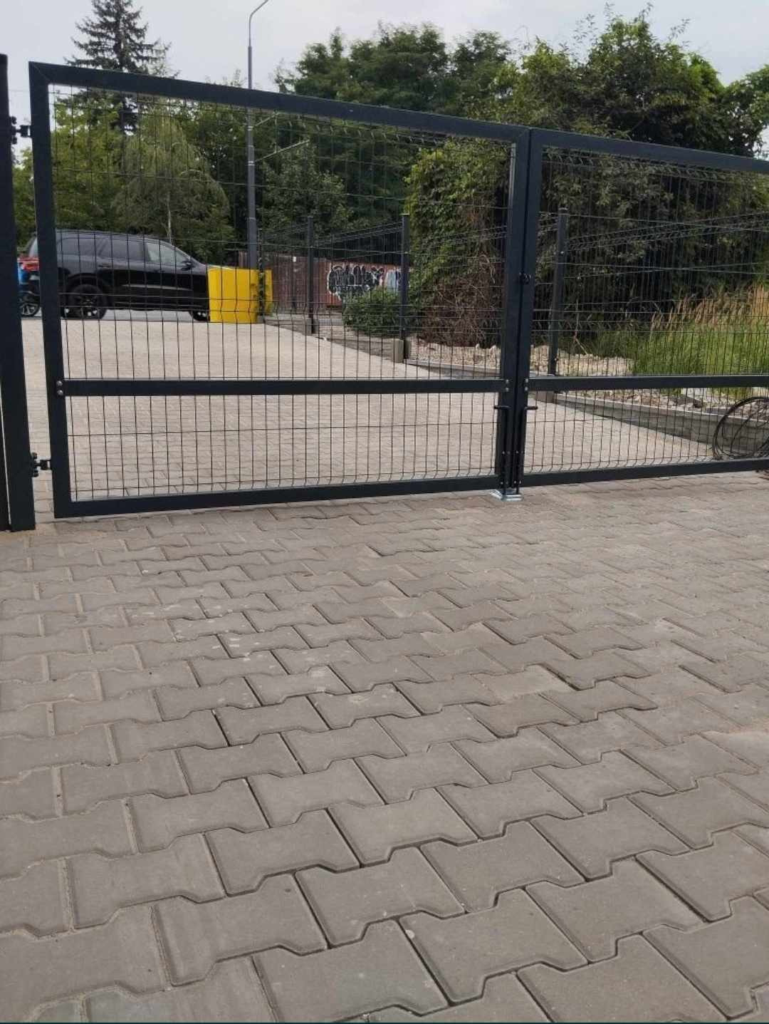 Otwarta, dwuskrzydłowa brama z metalowej siatki panelowej, widok z zewnątrz na szary chodnik z kostki brukowej i częściowo widoczny zaparkowany czarny samochód za bramą.