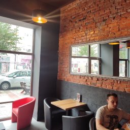 Horstbud - Wnętrze kawiarni z ceglaną ścianą, lustrem w ozdobnej ramie, drewnianymi stolikami i krzesłami w kolorze czarnym i czerwonym, oświetlone ciepłym światłem lamp wiszących.