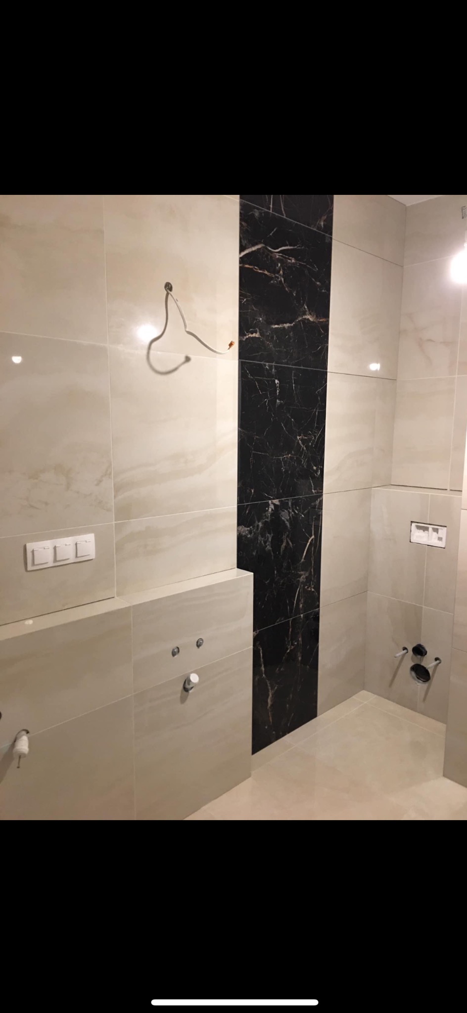 Łazienka w trakcie wykańczania, z beżowymi płytkami i pionowym pasem z czarnych płytek imitujących marmur; widoczne niedokończone instalacje wodno-kanalizacyjne i elektryczne.