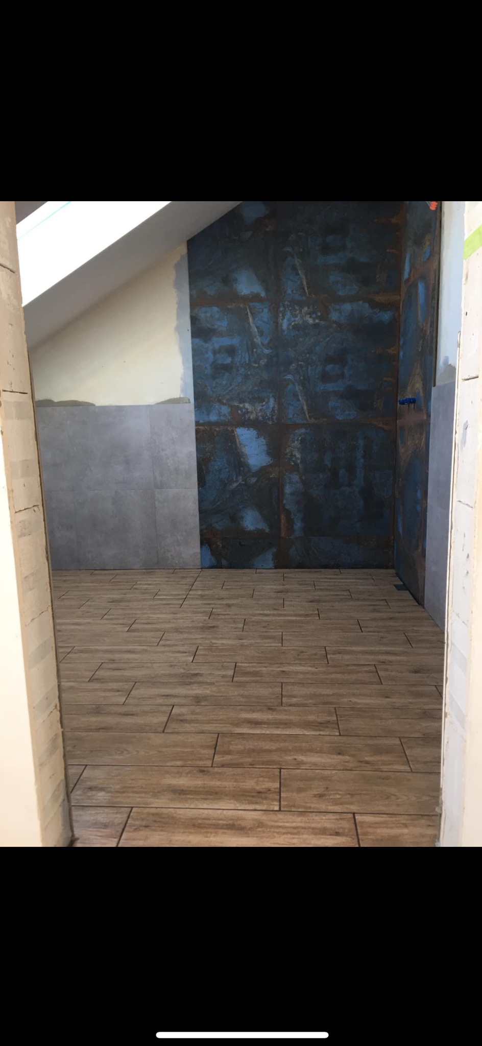 Łazienka na poddaszu w trakcie wykańczania: drewnopodobne płytki na podłodze, częściowo wyłożone szare i niebiesko-rdzawe płytki na ścianach, widoczne niedokończone połączenia i skos dachu.