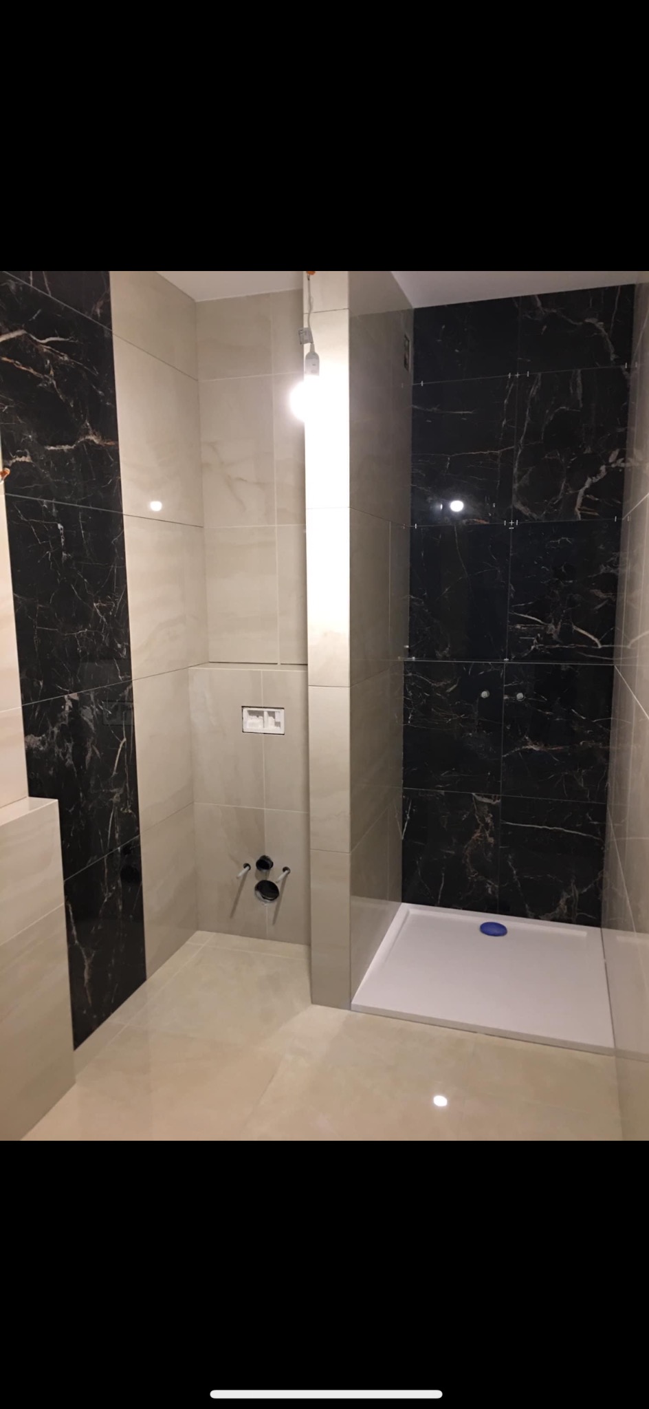 Wykończona łazienka z czarno-białymi płytkami imitującymi marmur na ścianach, biały brodzik z niebieskim korkiem, widoczne przyłącza wodne do baterii.