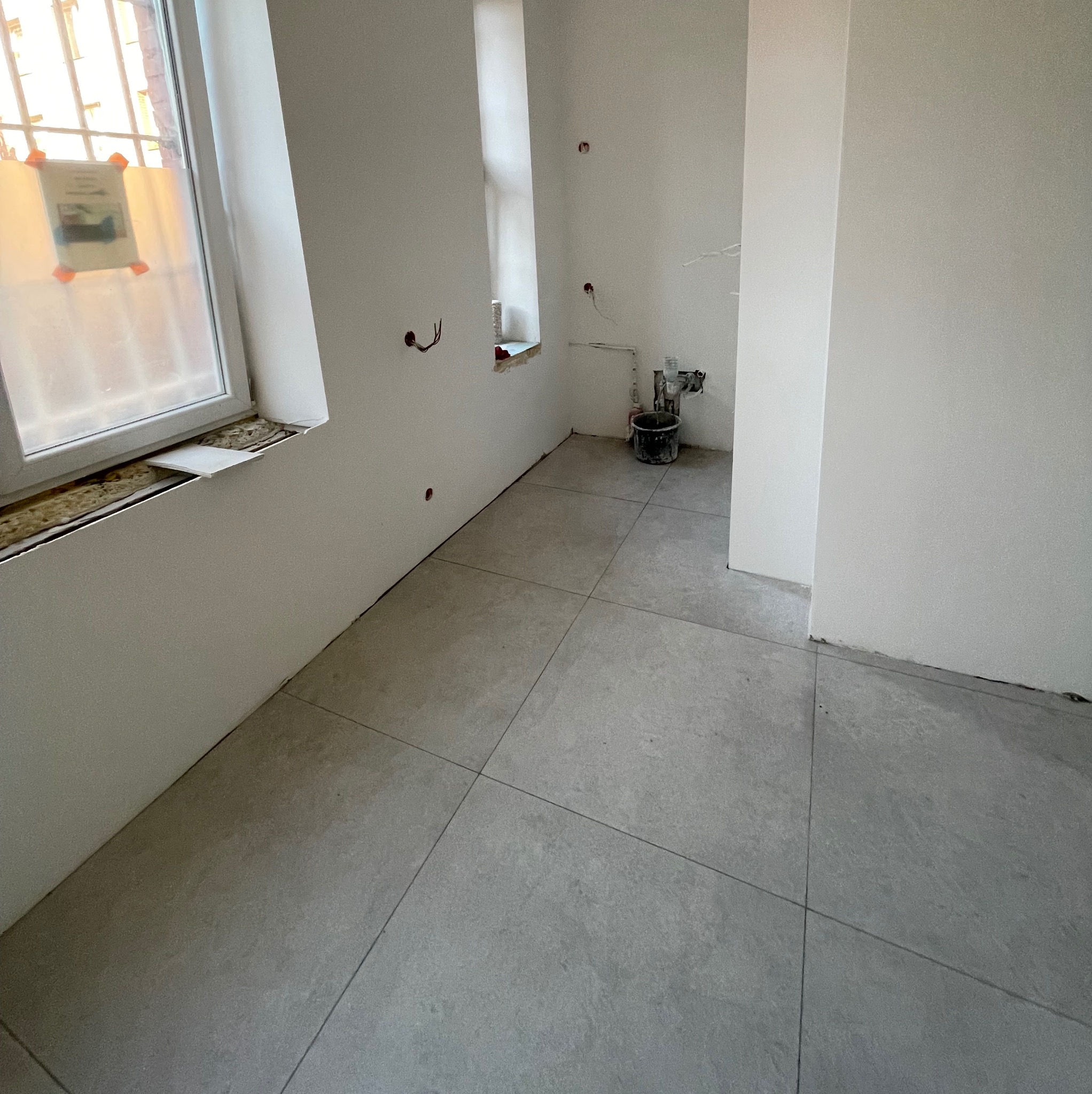 Szare płytki podłogowe ułożone w kwadraty w surowym wnętrzu łazienki w trakcie remontu, widoczne instalacje elektryczne i hydrauliczne, okno z zabezpieczeniem.